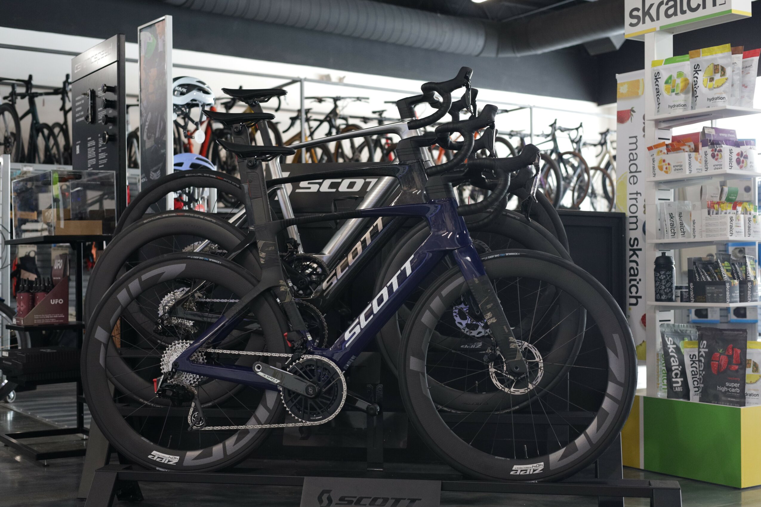Two black and blue Scott bicycles displayed in a store, with other bikes and shelves in the background.