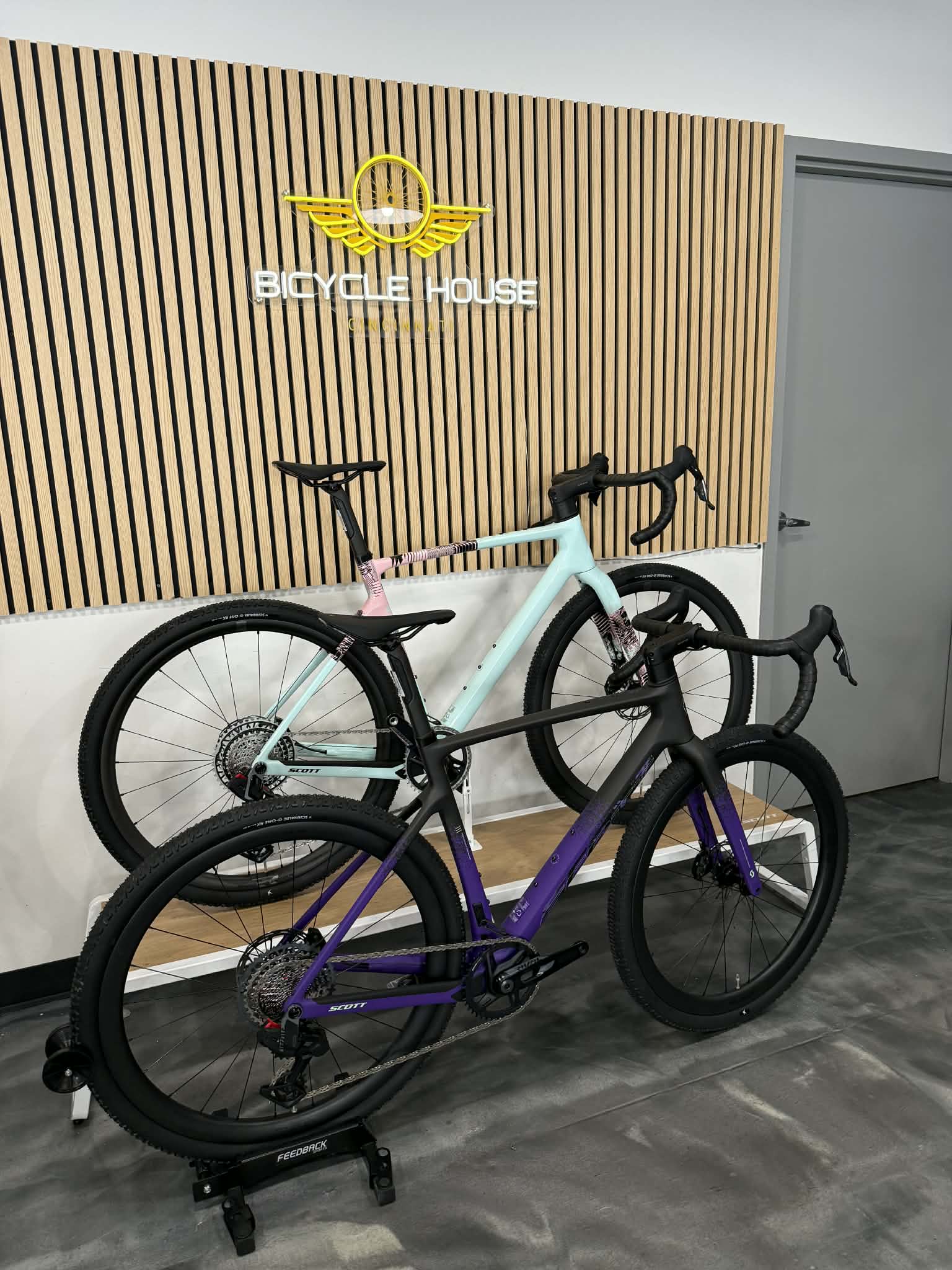 Two bicycles, one pink and white, and one purple and black, are displayed indoors against a wooden wall with a logo.