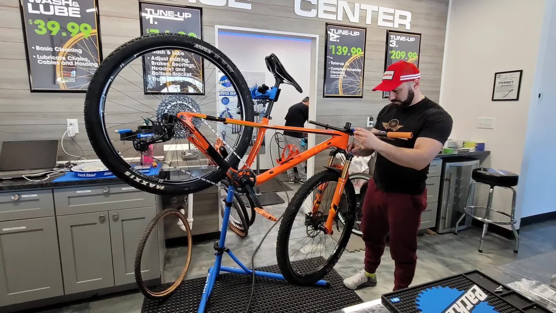 Person in a red cap and black shirt inspecting an orange mountain bike inside a store. Store signs and counters are visible.