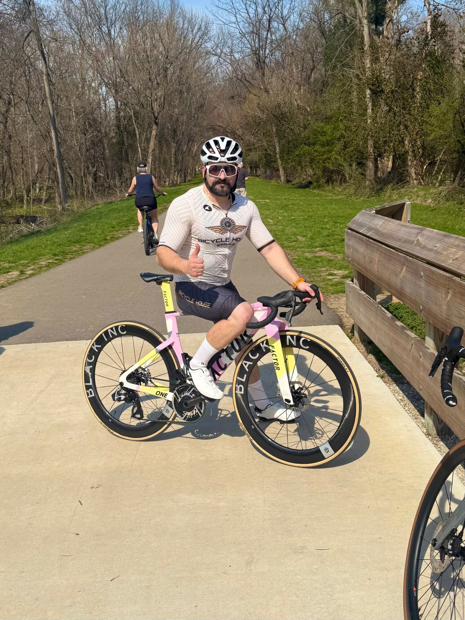Person wearing a helmet and sunglasses, sitting on a pink and black bicycle on a paved path in a park, with trees in the background.