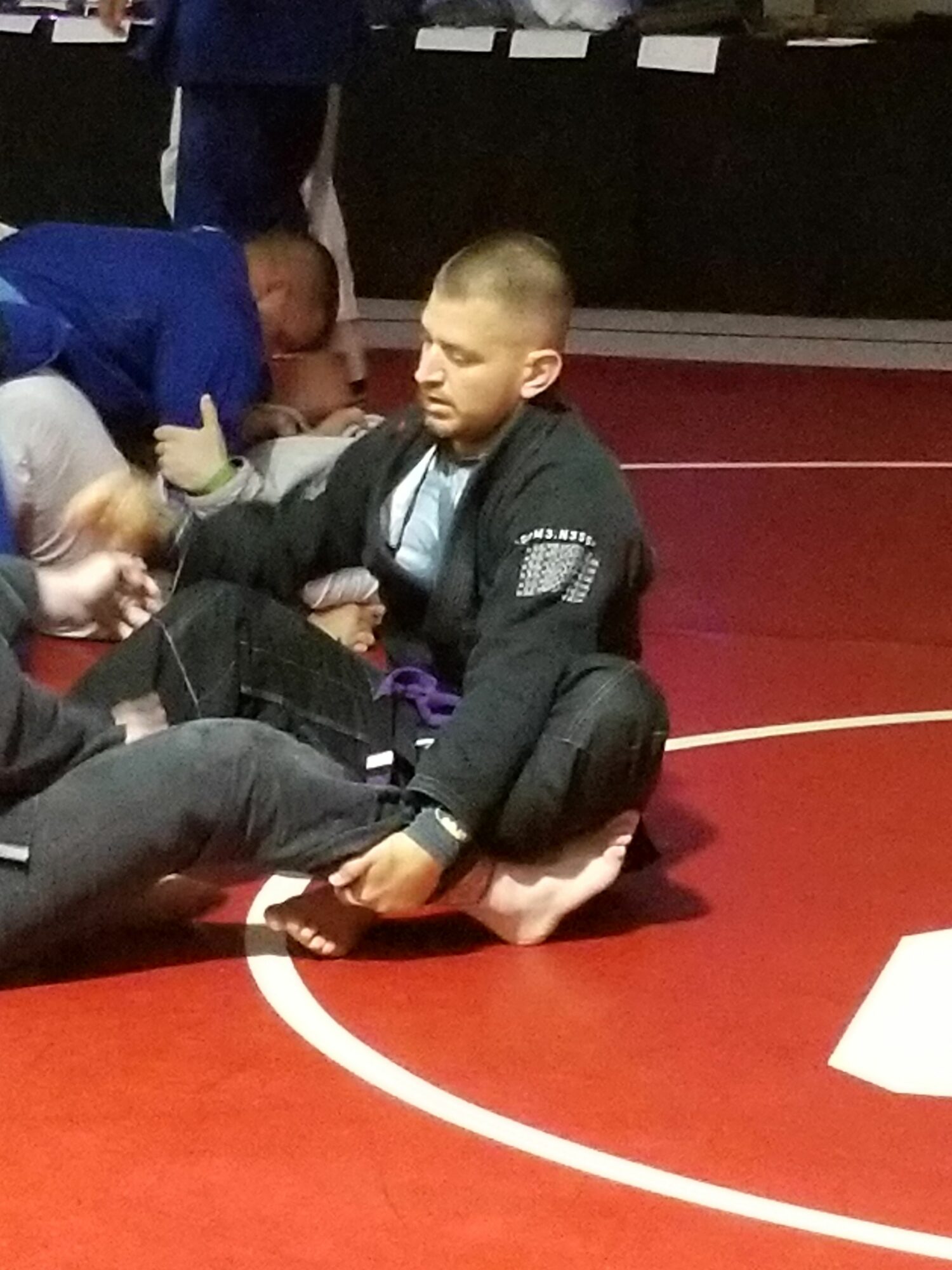 Two children on a red mat, one sitting and the other lying down, in a gym or sports facility.