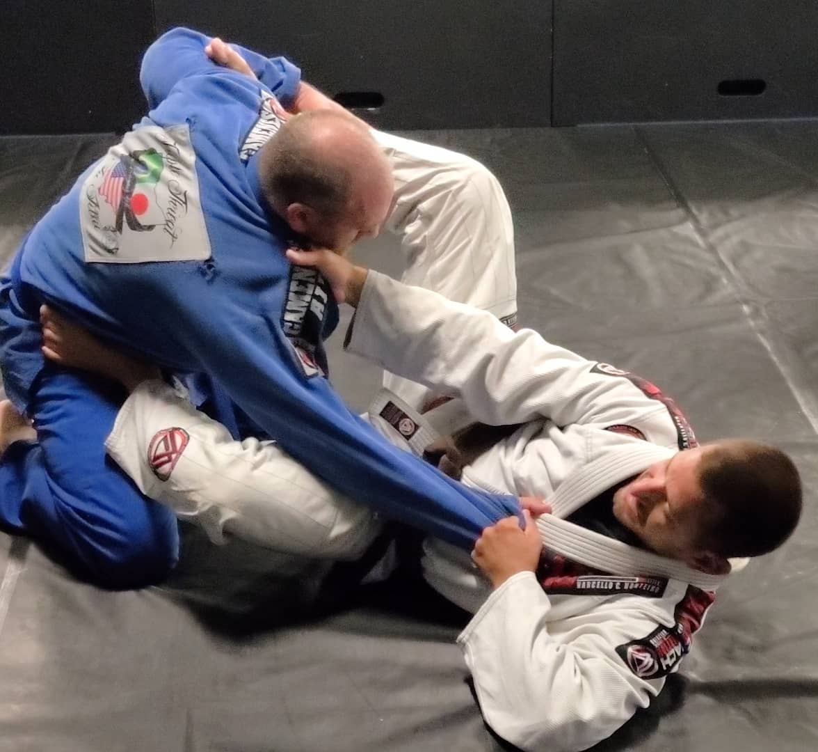 Two people practicing Brazilian Jiu-Jitsu on a mat, one in a blue gi and the other in a white gi, grappling.