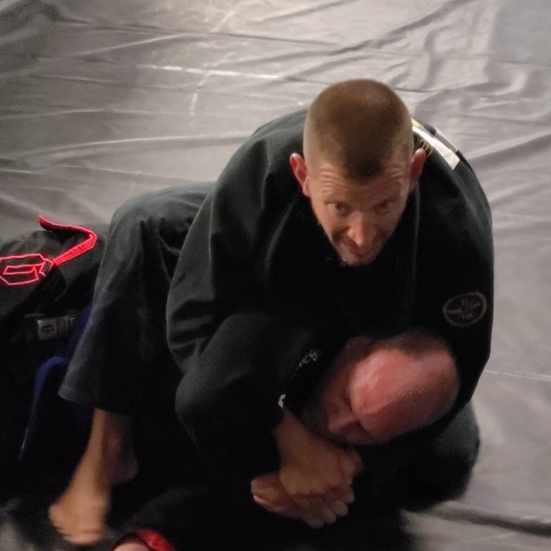 Two people practicing martial arts on a mat, one on top of the other, wearing black uniforms.