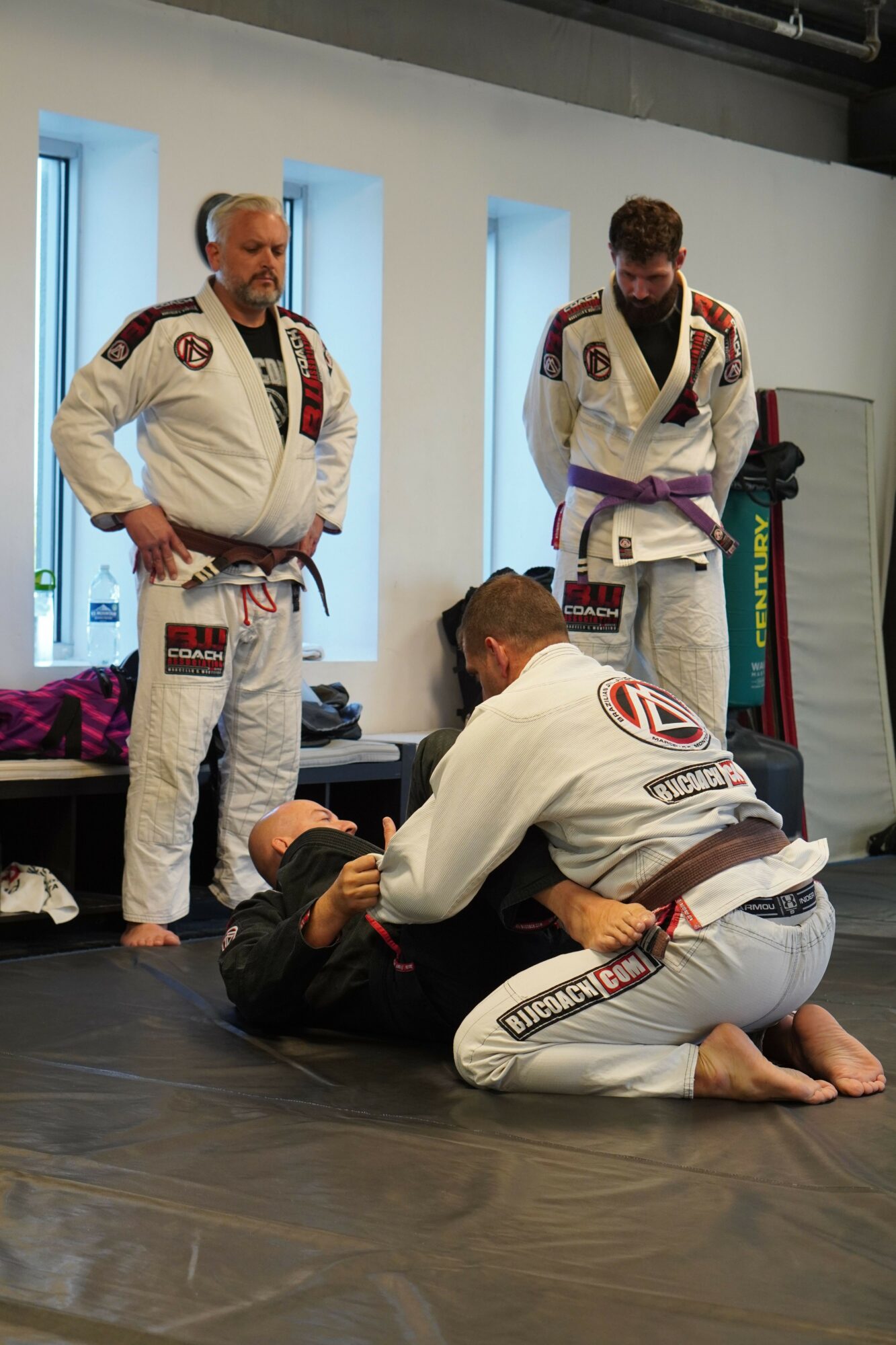 Two people in Brazilian Jiu-Jitsu gis practicing on the floor, with a woman standing nearby observing, in a room with large windows.