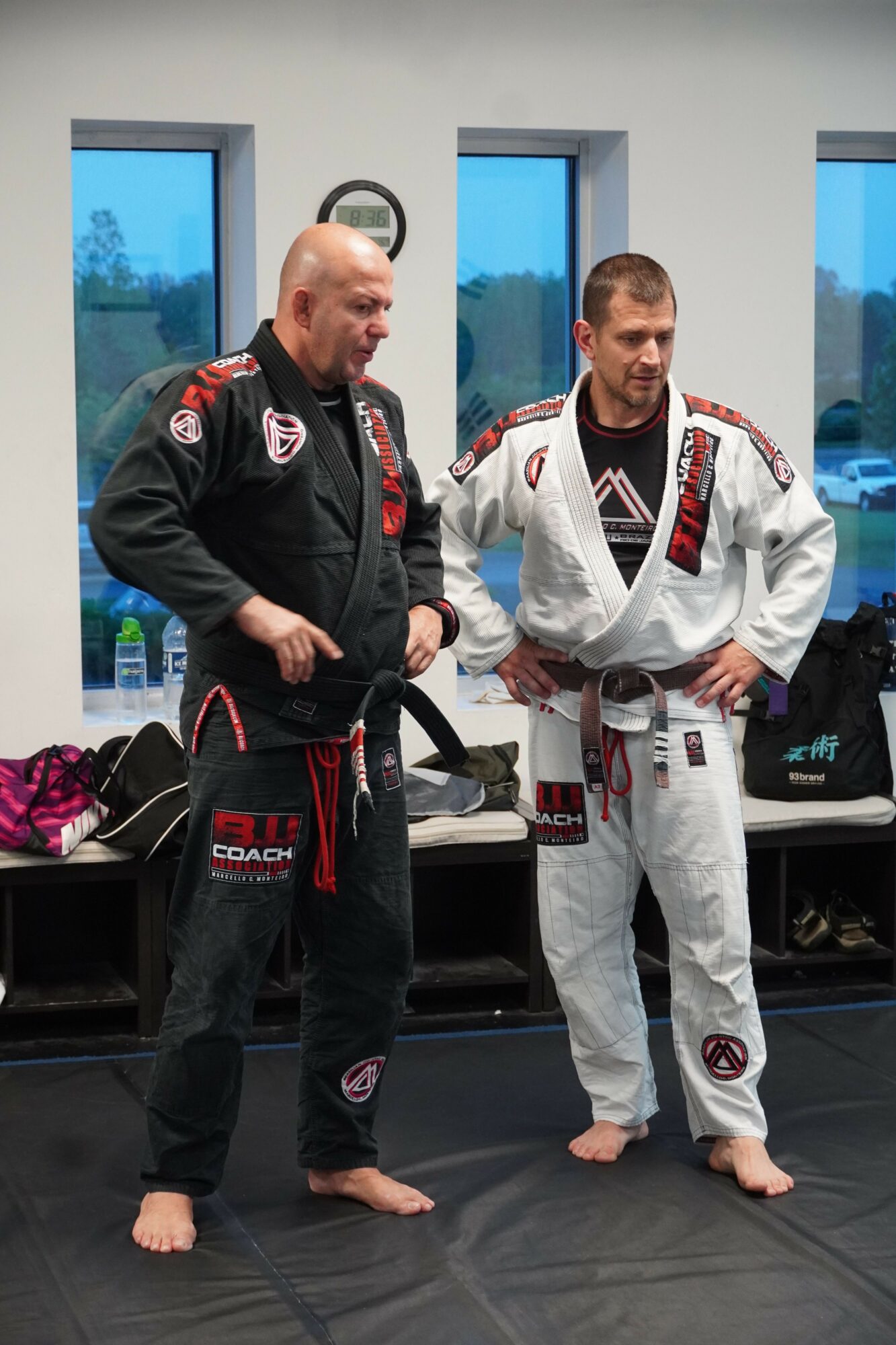 Two people in martial arts uniforms standing in a room with windows and a television, one with a black gi and the other with a white gi.
