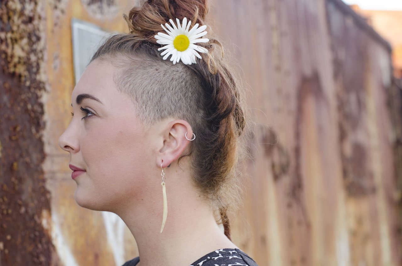 Side profile of a woman with a daisy in her hair, earrings, and a patterned top, standing outdoors.