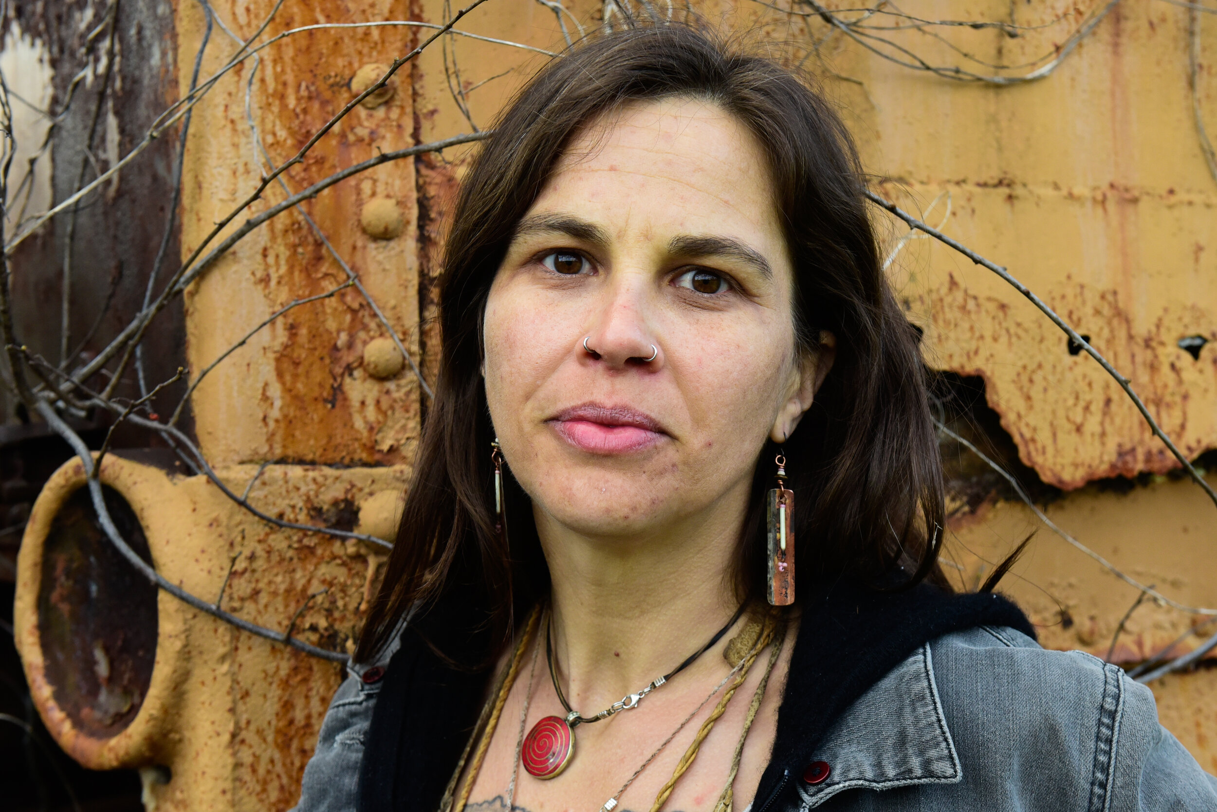 Woman with dark hair and earrings outdoors, background of rusted metal and branches, wearing a denim jacket.
