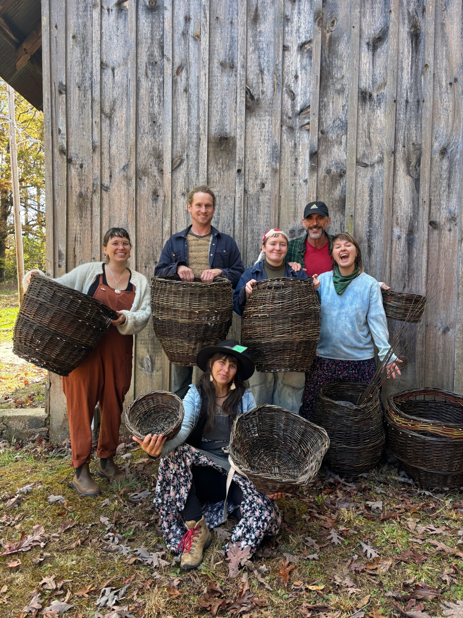 Six people outdoors in front of a wooden fence, holding baskets, smiling, with trees in the background.