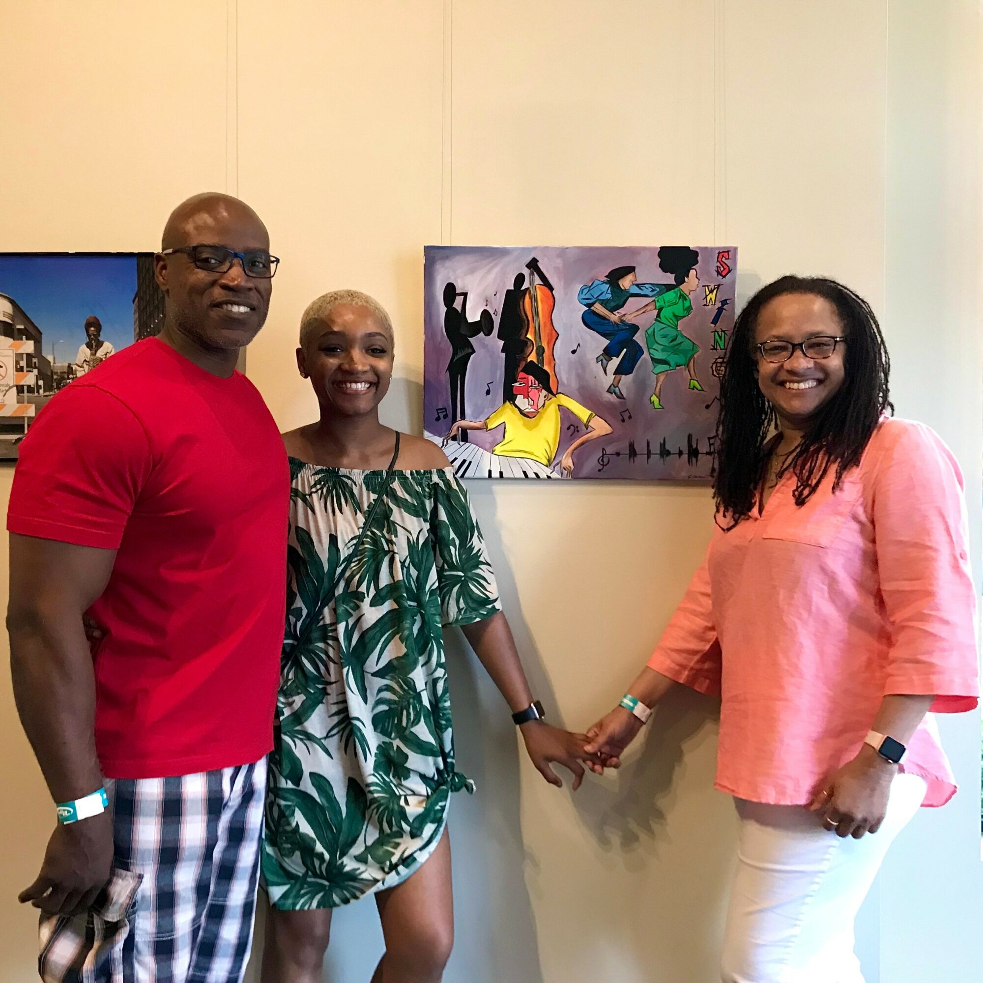 Three people standing in front of a colorful painting, holding hands, smiling, in a gallery setting.