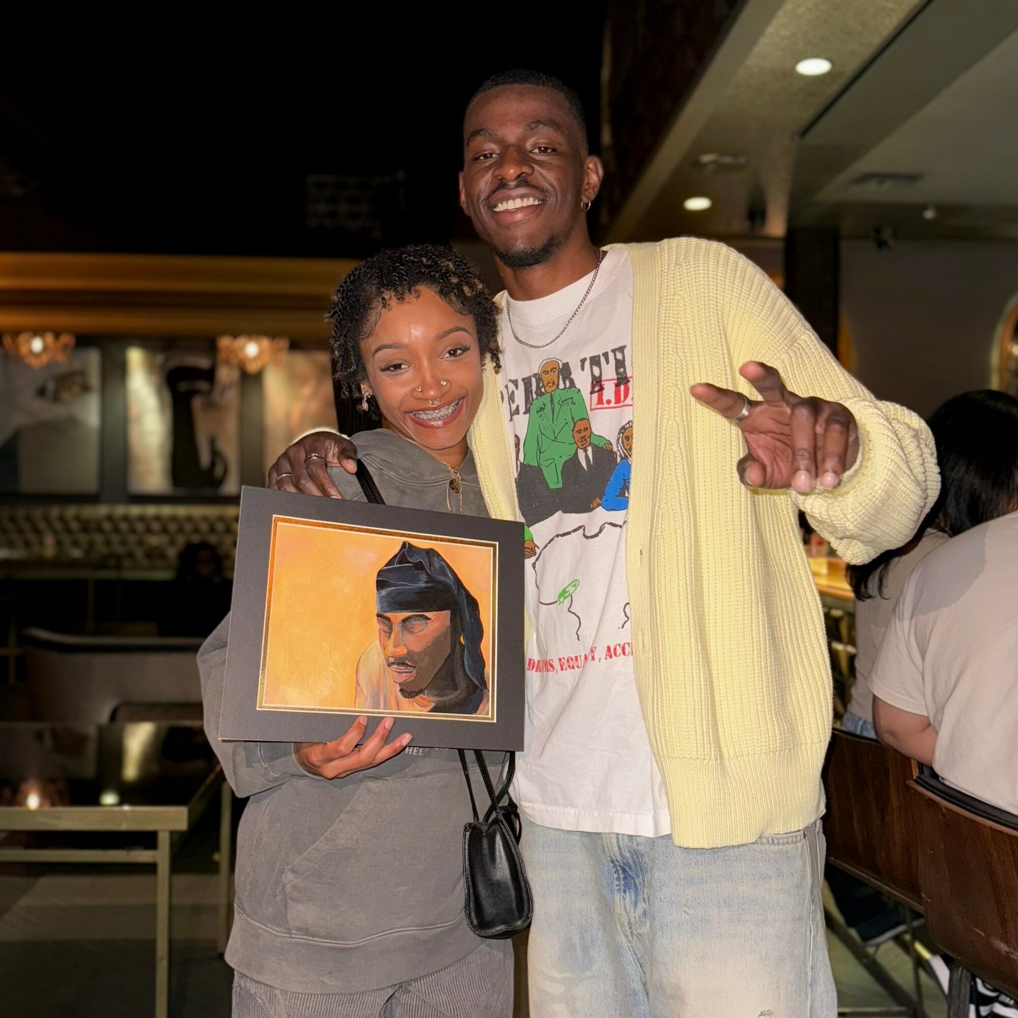 Two people smiling, one holding a framed painting of a woman with a headscarf, in a restaurant setting.