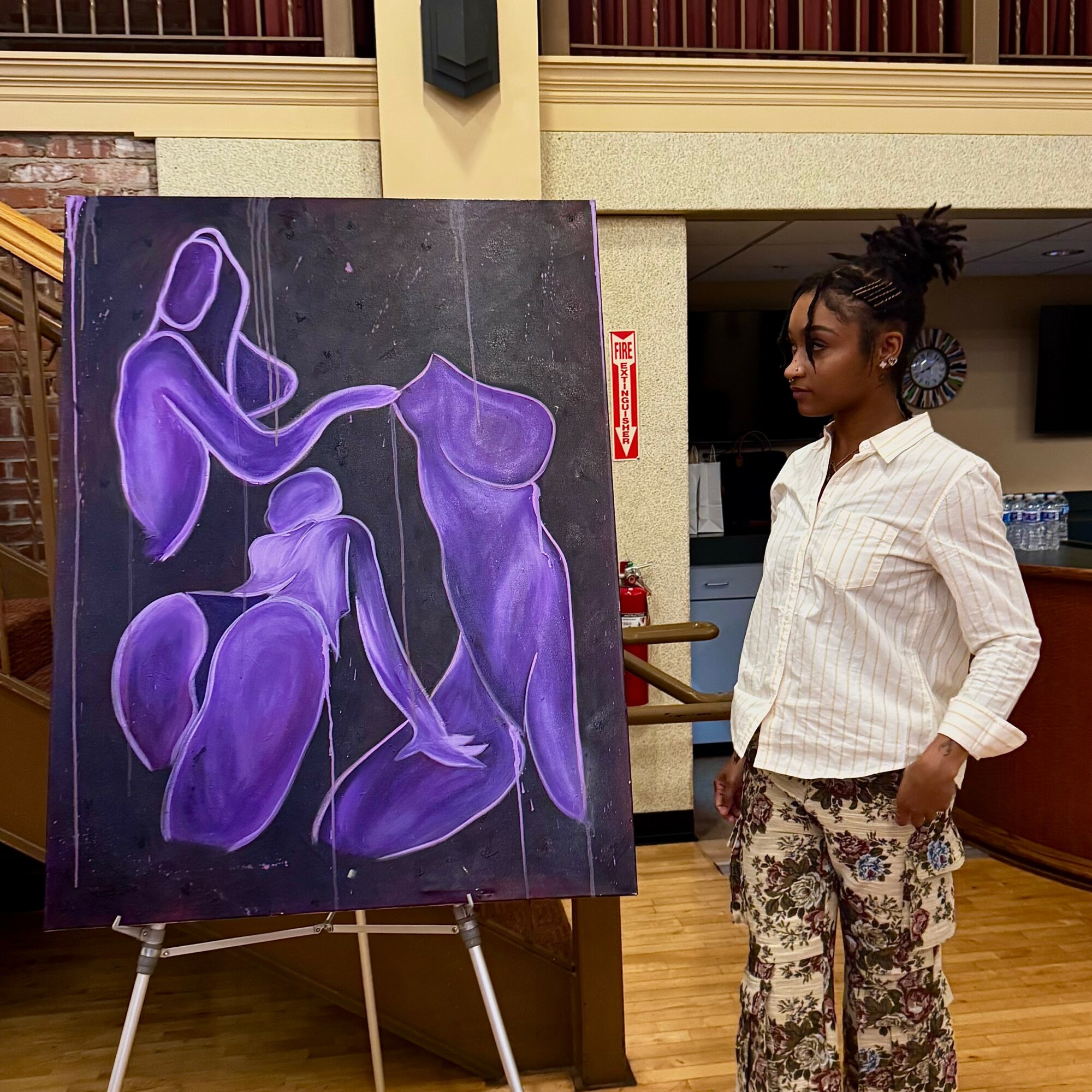 Young woman with braided hair, wearing a white shirt and patterned pants, stands indoors near a purple abstract painting.