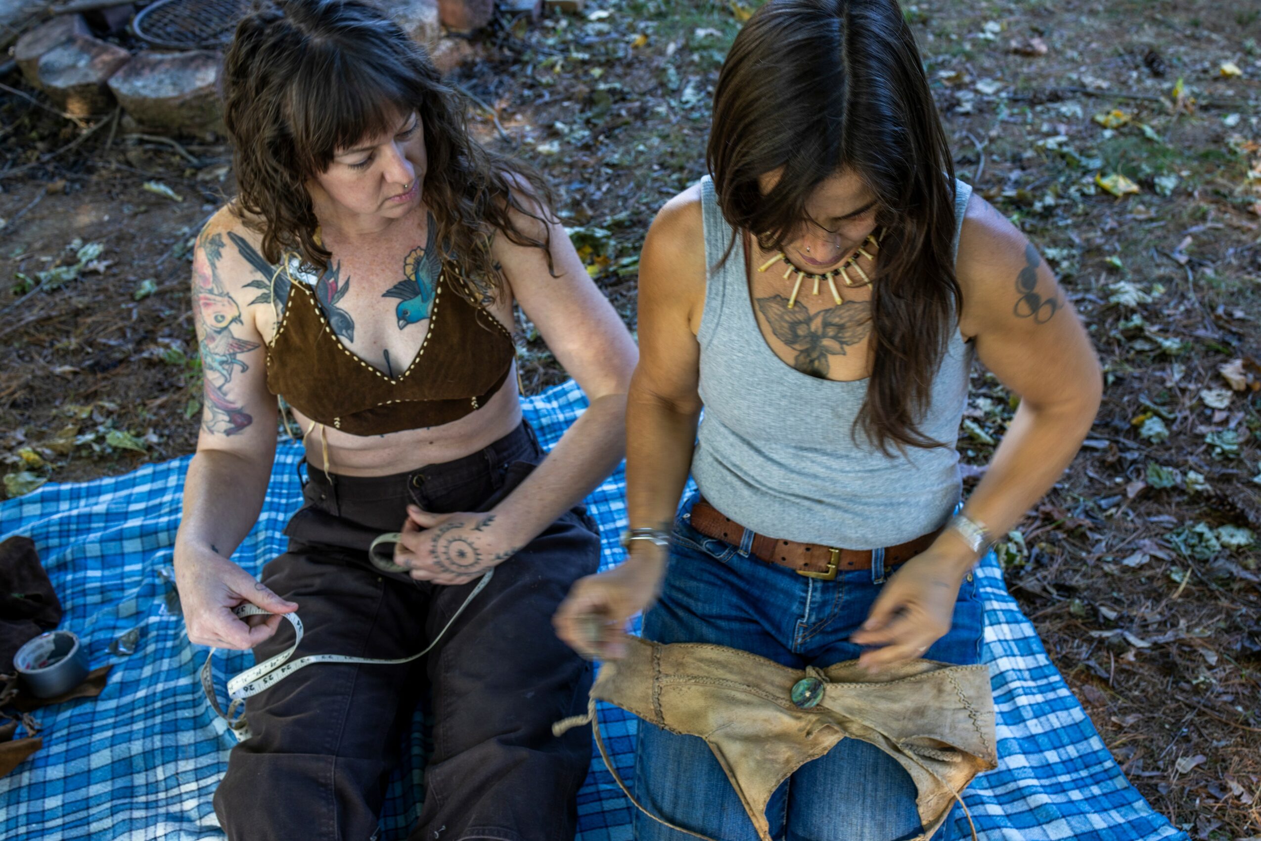 Two women sit on a blue checkered blanket outdoors, one with long dark hair and tattoos, the other with curly hair, holding a bag.