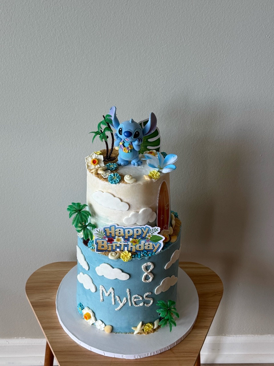 Two-tier birthday cake with blue and white clouds, a tiger figure on top, and tropical decorations, with 'Happy Birthday Myles' written.