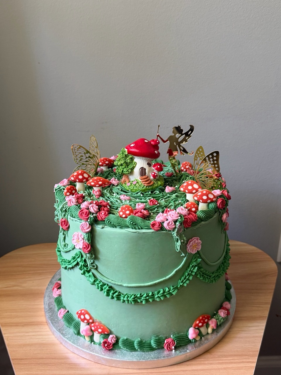 Two-tiered green cake decorated with pink and red flowers, mushrooms, gold butterflies, and a small figure on top, on a wooden table.