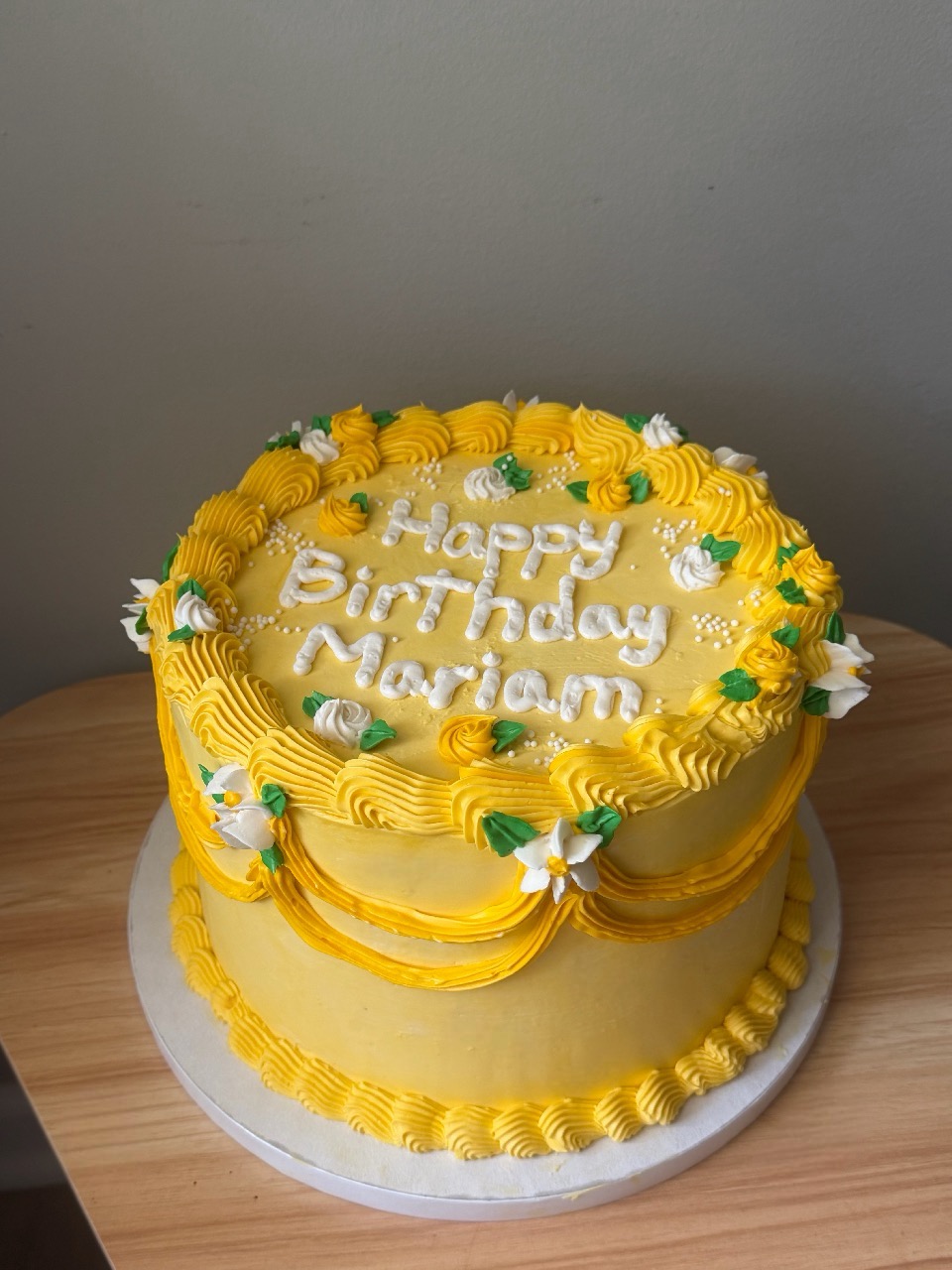 Round yellow birthday cake with white and green decorations, message on top, on a wooden surface, gray background.