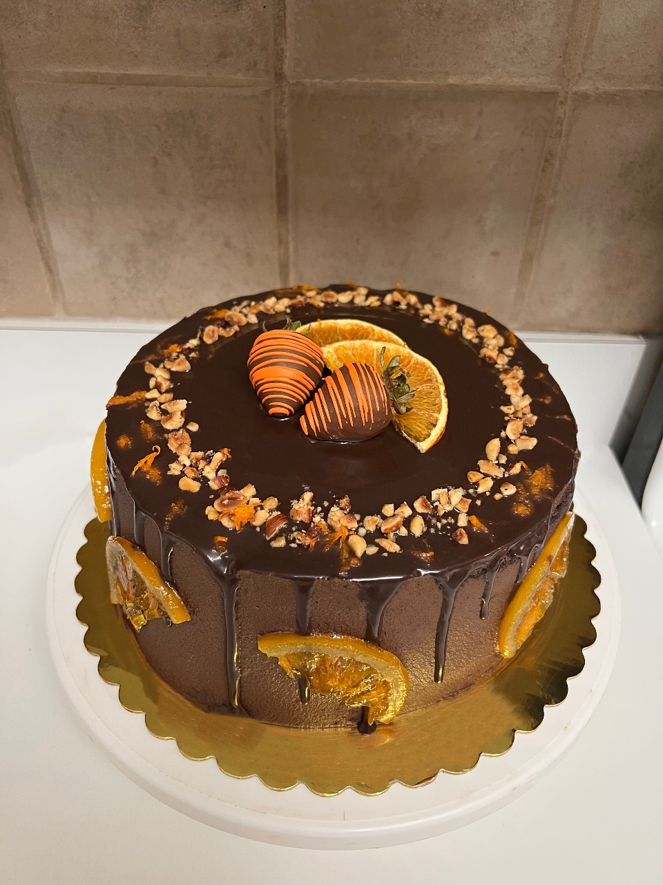 Chocolate cake with orange slices and chocolate decorations on top, decorated with nuts around the edge, on a white surface.