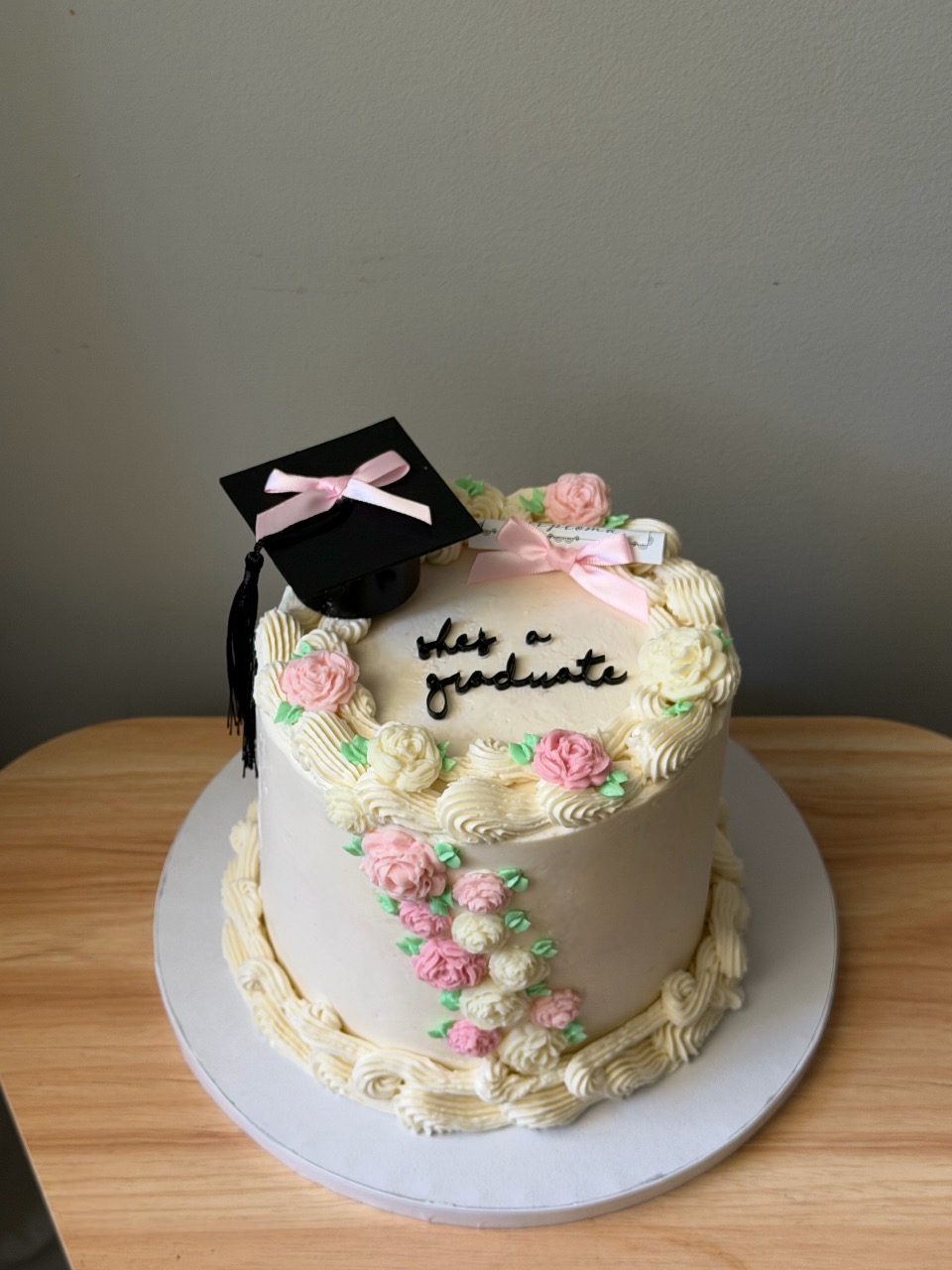A graduation cake decorated with pink and white flowers, a black graduation cap with a pink ribbon, on a white base.