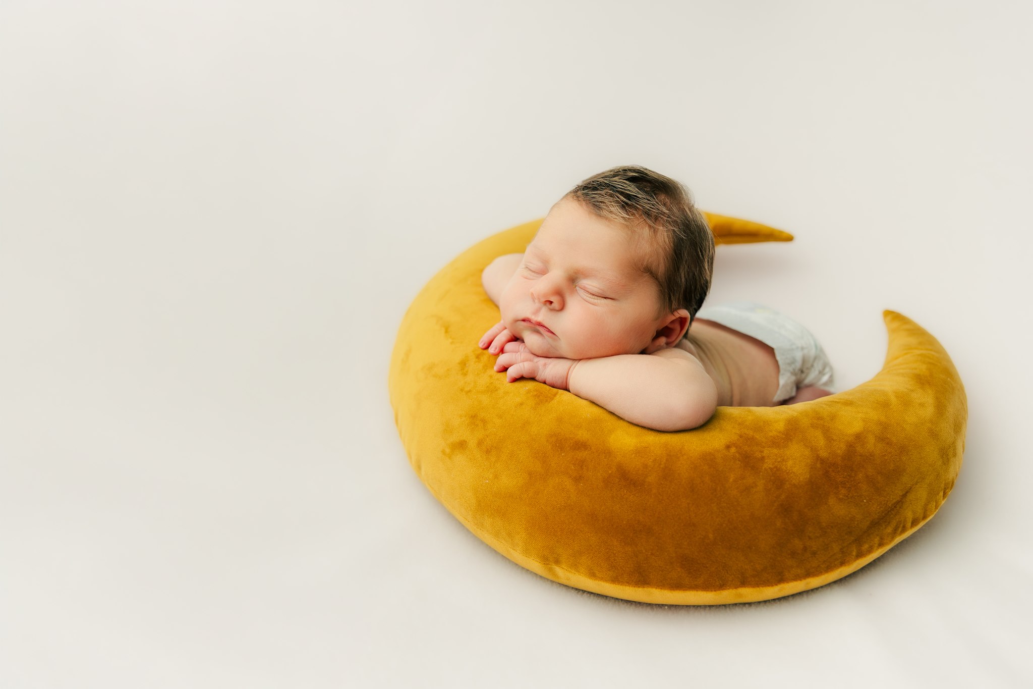 Baby sleeping on a yellow crescent-shaped pillow, resting with hands under chin, against a plain white background.