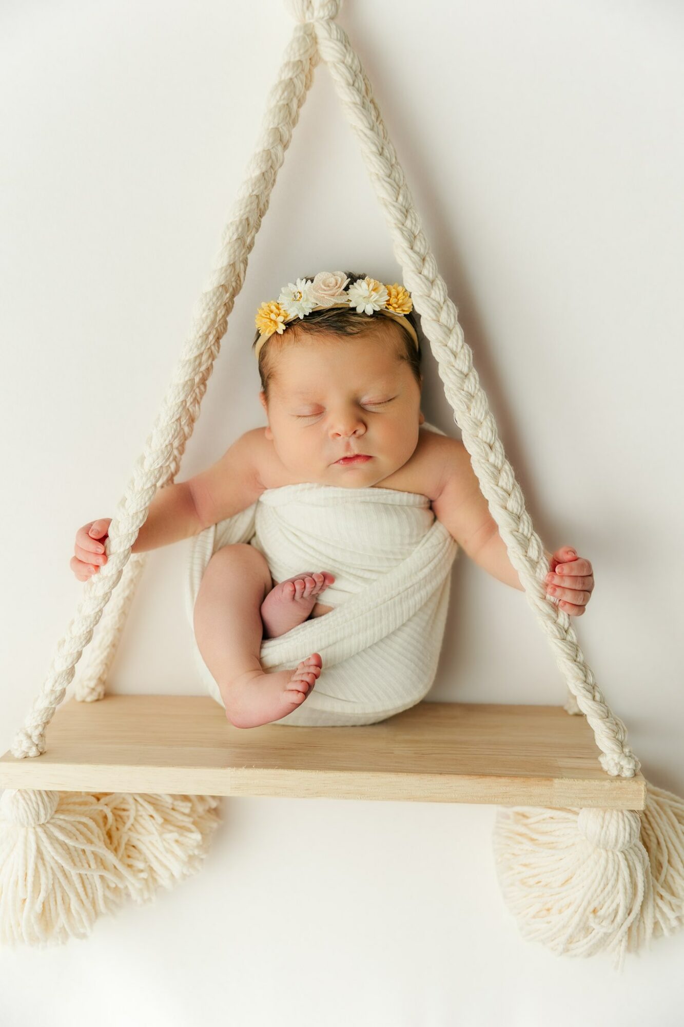 Baby wrapped in a white blanket, sleeping on a wooden swing with a floral headband, against a plain background.