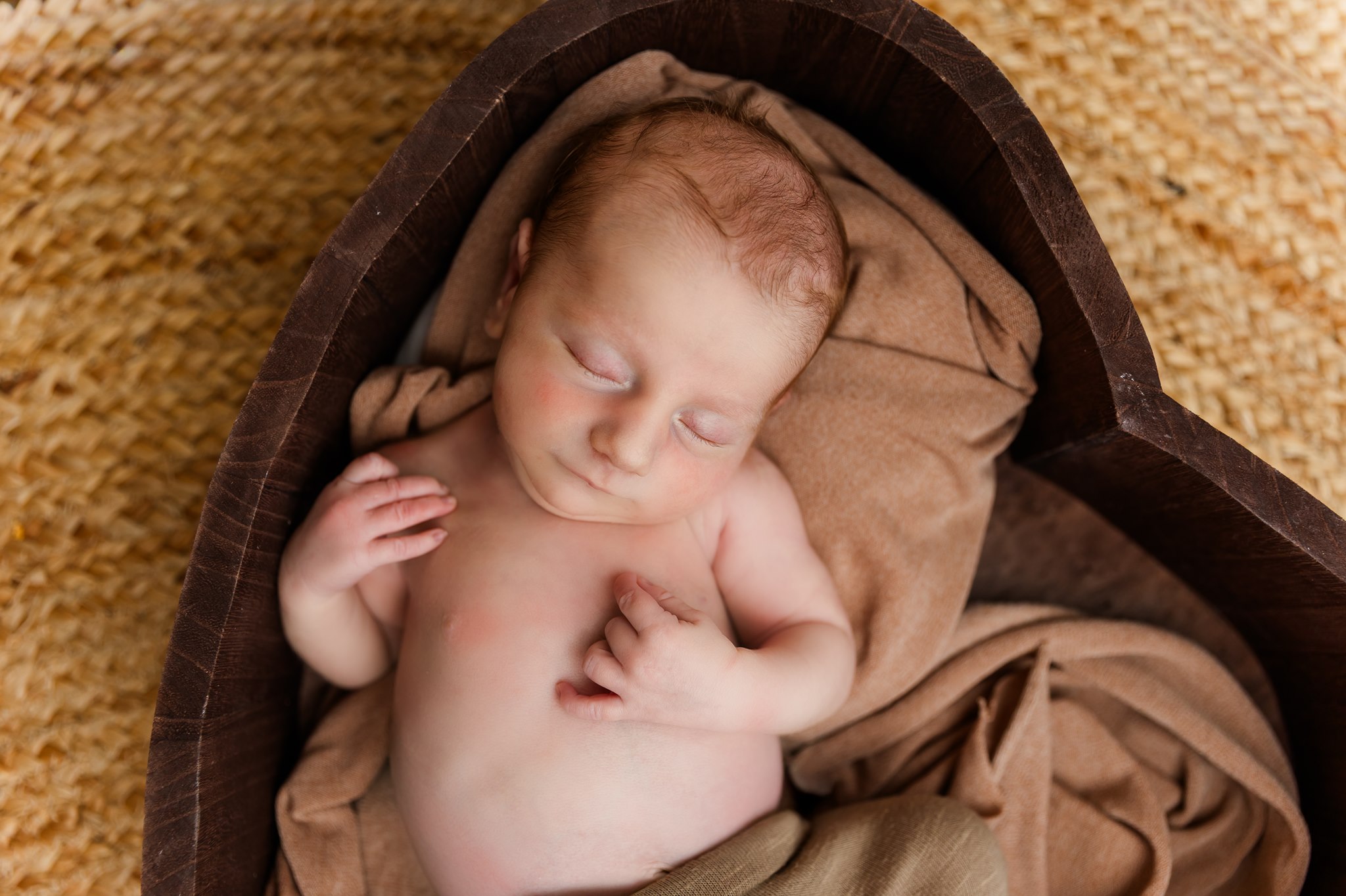 Newborn baby sleeping on a blanket inside a wooden heart-shaped frame, with a beige cloth beneath.