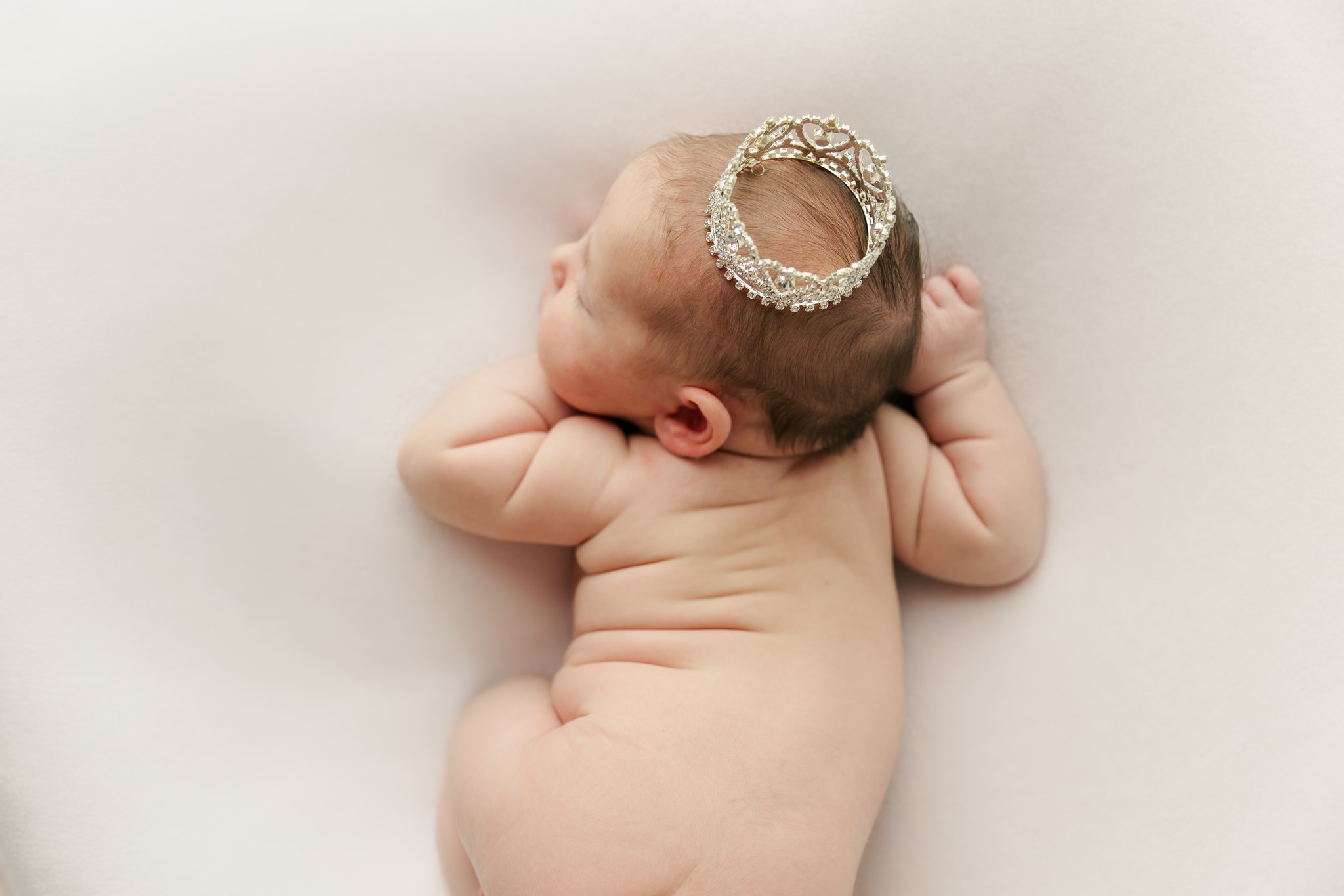 Baby lying on side with head resting on arm, wearing a decorative headband, on a white surface.