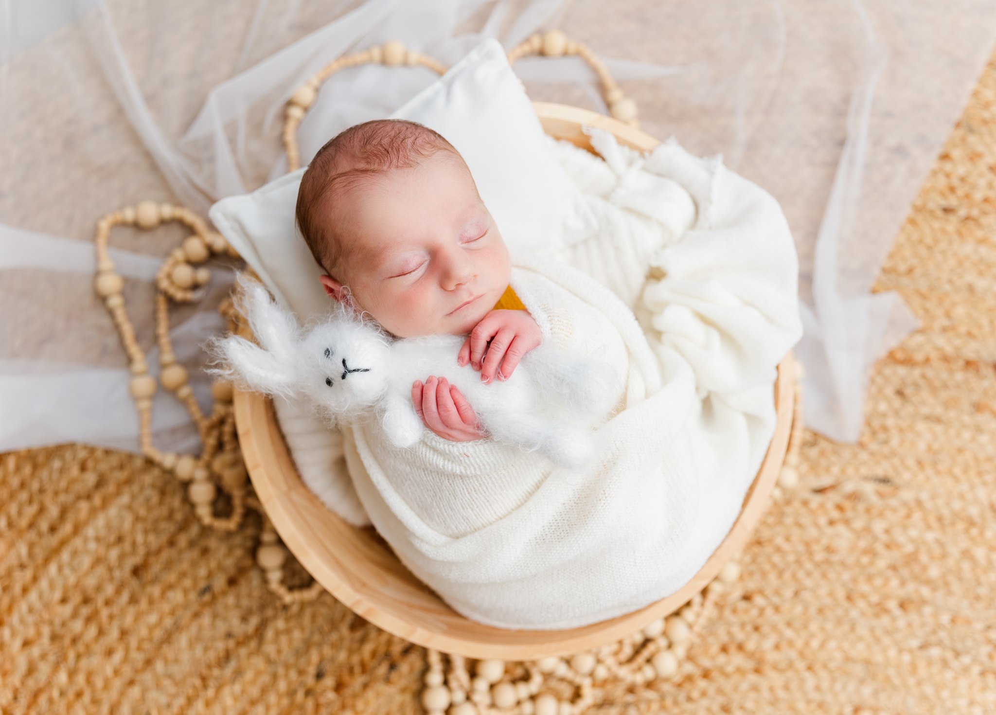 Newborn baby sleeping in a basket, holding a small white stuffed bunny, wrapped in a white blanket, on a textured surface.