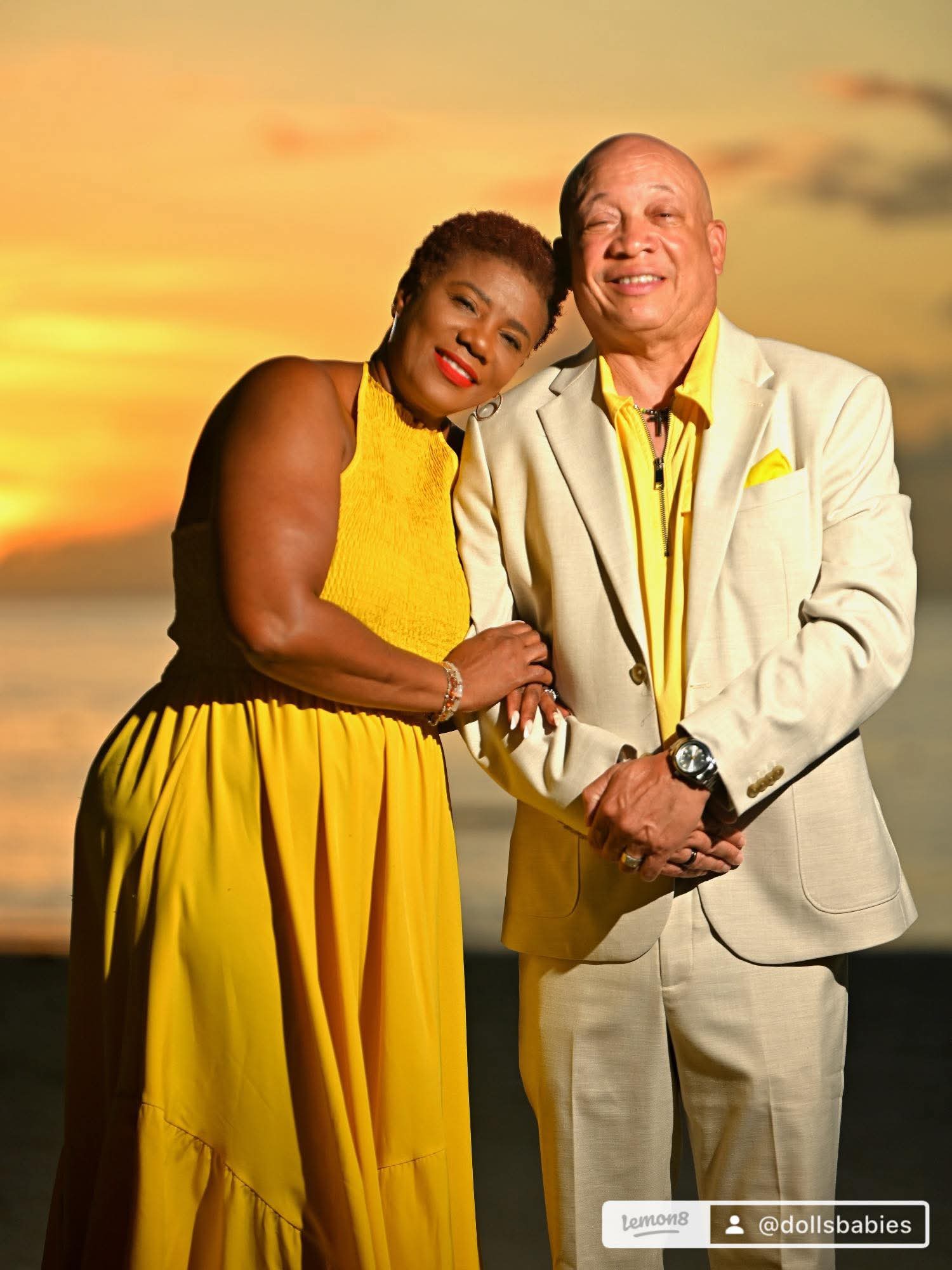 A woman and a man stand close together outdoors at sunset, both smiling, with the woman resting her head on the man's shoulder.