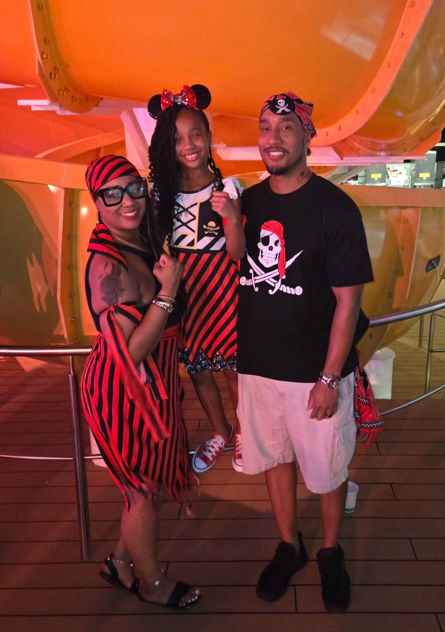 Three people standing together on a wooden deck, smiling, with colorful structures in the background, wearing casual and themed clothing.