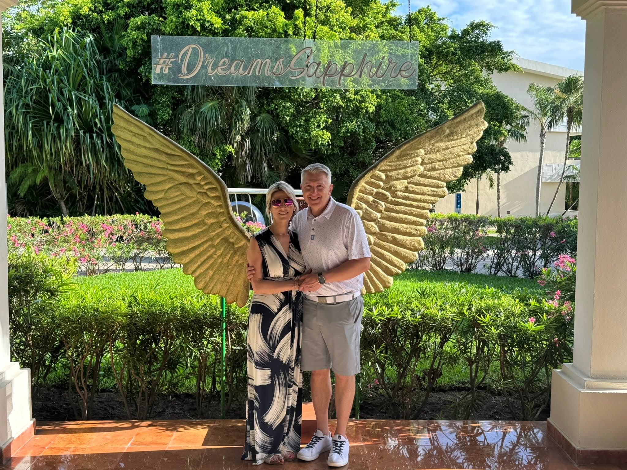 Two people stand in front of a decorative winged sculpture outdoors, holding hands, with greenery and a building in the background.