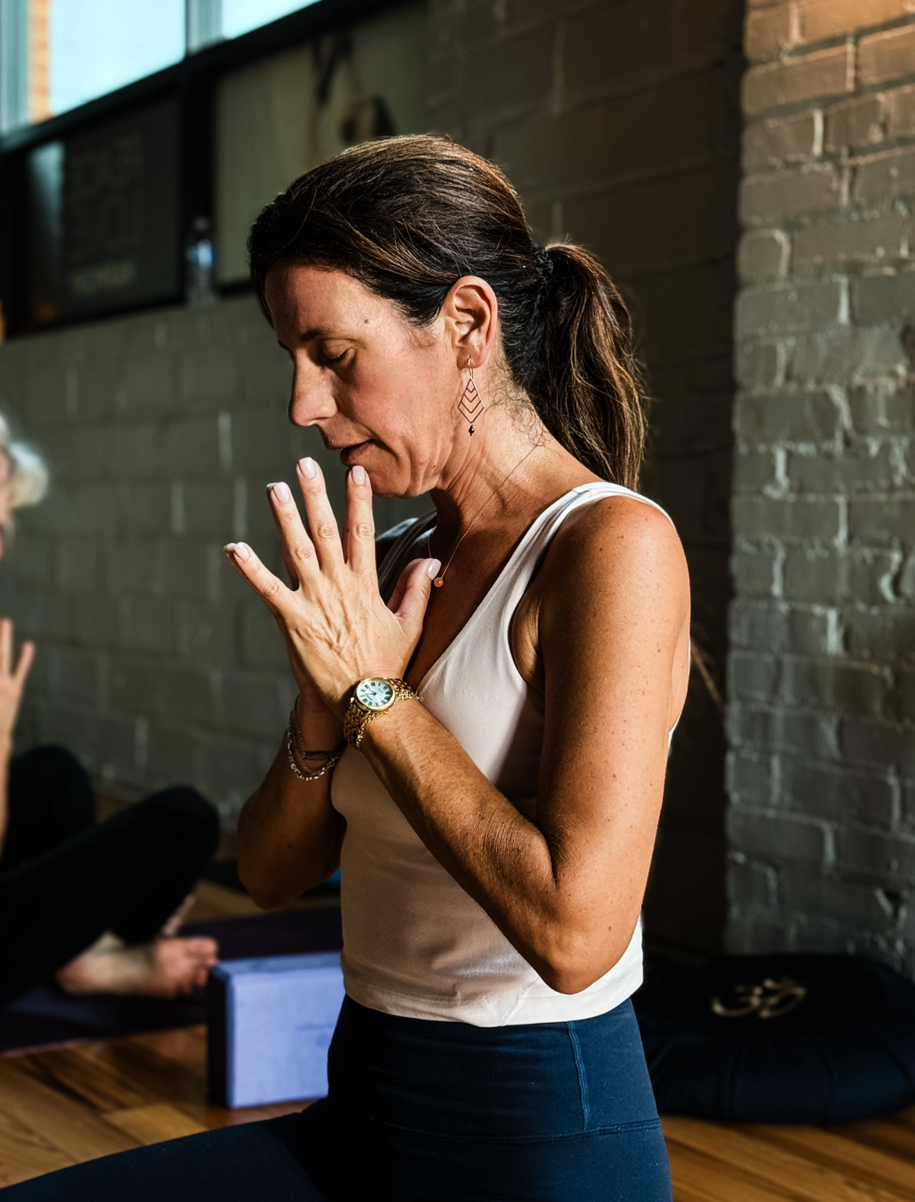 Woman with brown hair in a ponytail, wearing a white sleeveless top and dark skirt, praying with hands together, eyes closed.