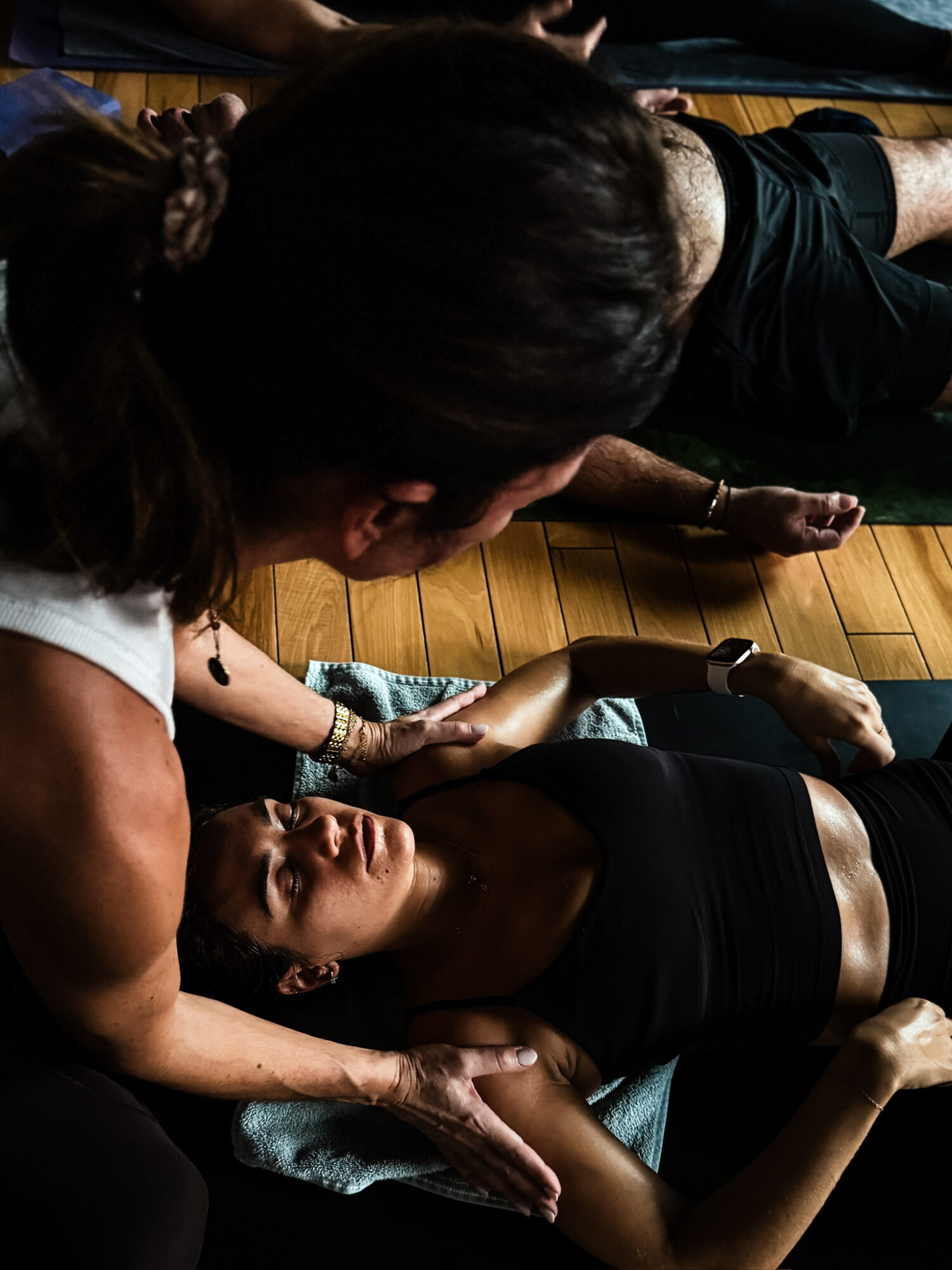Two women practicing yoga on mats, one lying down and the other assisting, in a wooden-floored room.