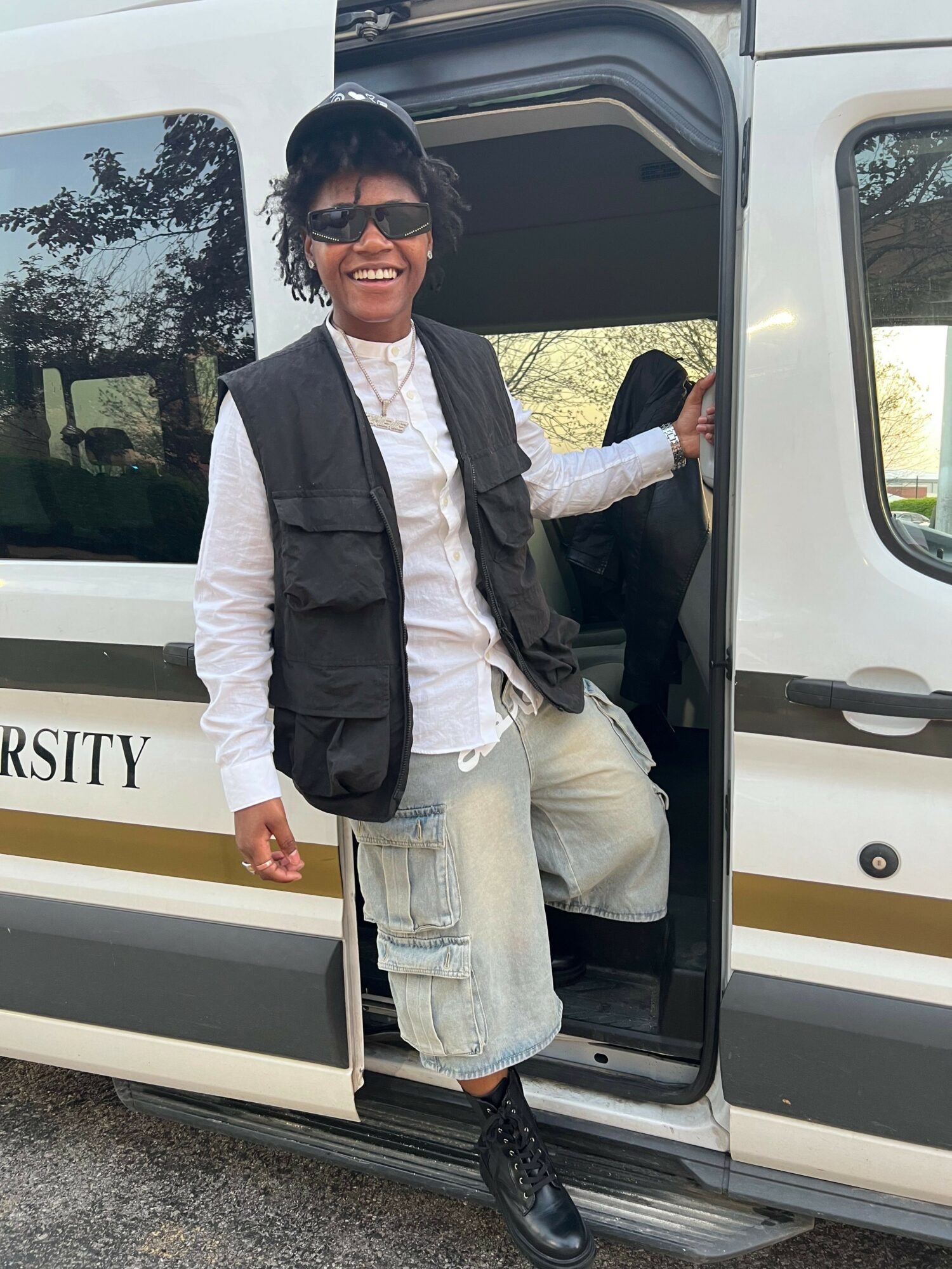 Person standing outside a vehicle, smiling, wearing sunglasses, a black jacket, white shirt, and light-colored pants.