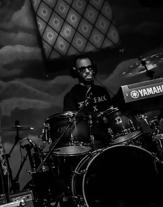 Person playing drums on stage with a Yamaha keyboard, dark background, and patterned backdrop, in black and white.