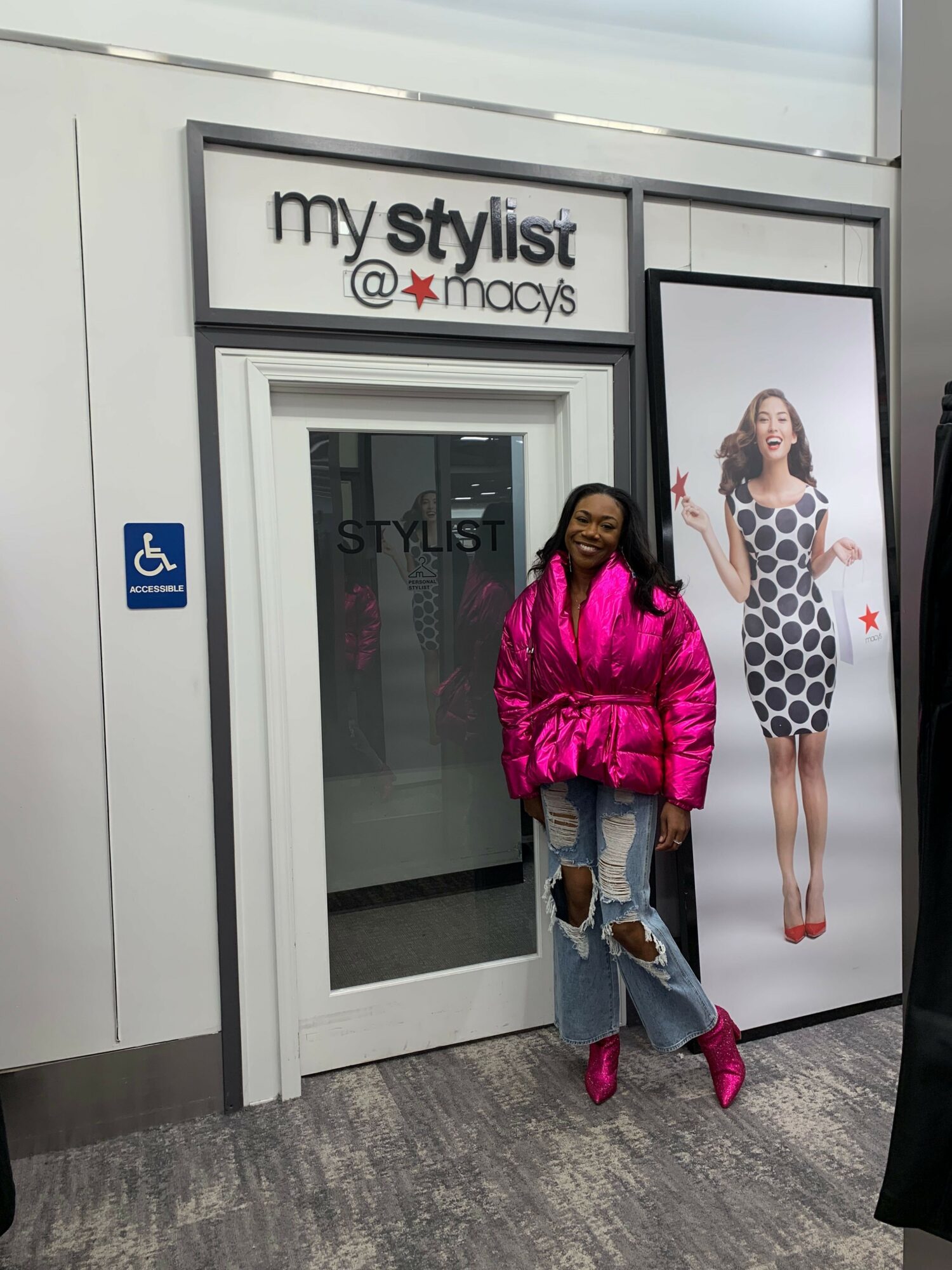 Woman in pink jacket and ripped jeans standing outside a store with a large poster of a woman in a polka dot dress.