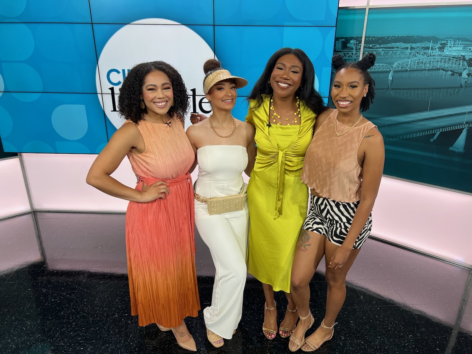 Four women standing together in a studio, smiling, with a blue background and a large circular logo.