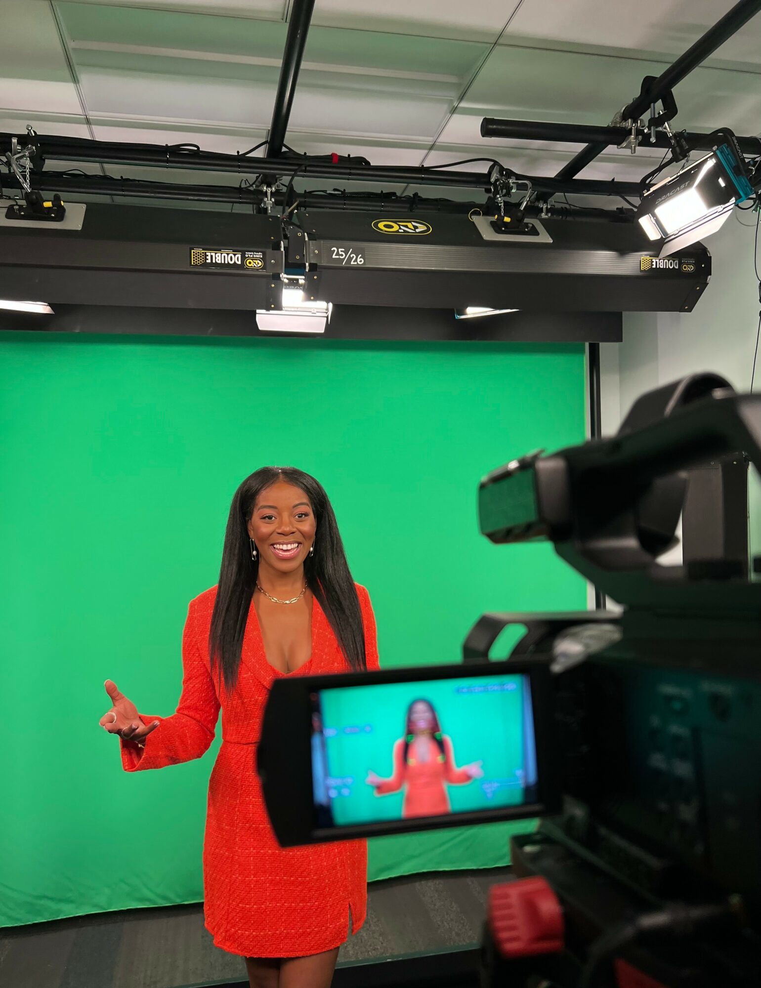Woman in red dress standing in front of green screen, smiling, with camera recording her, studio lighting overhead, 20 words
