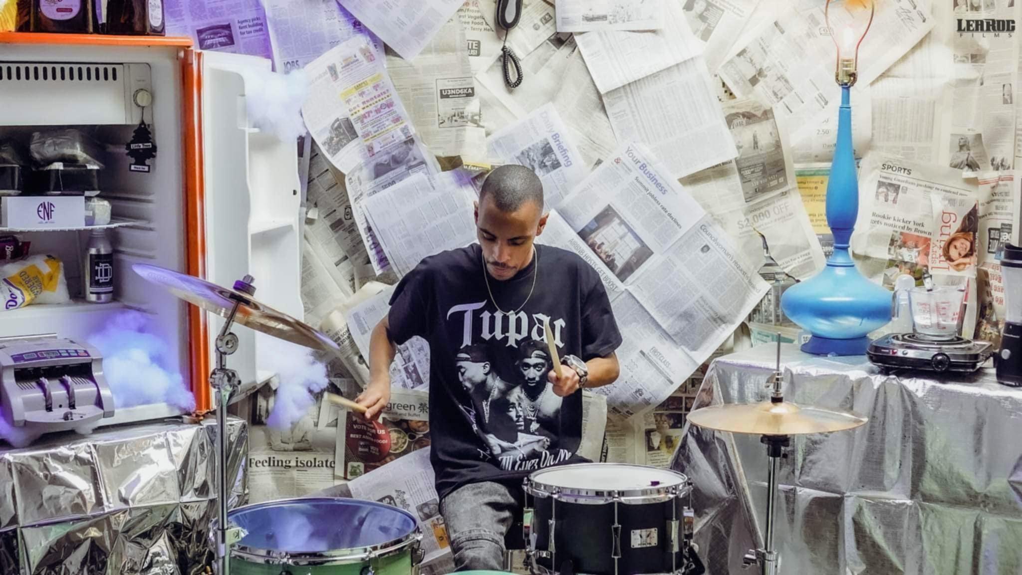 Person playing drums in a cluttered room with newspaper-covered walls and various objects, including a blue vase and a refrigerator.