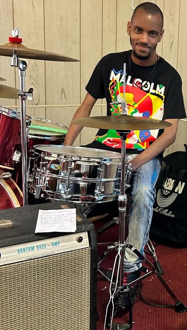Young man sitting behind a drum set, smiling, wearing a black T-shirt with colorful graphics, in a room with wooden walls.