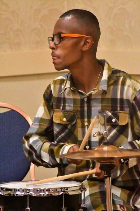 Person with glasses playing drums, wearing a plaid shirt, in a room with beige walls.