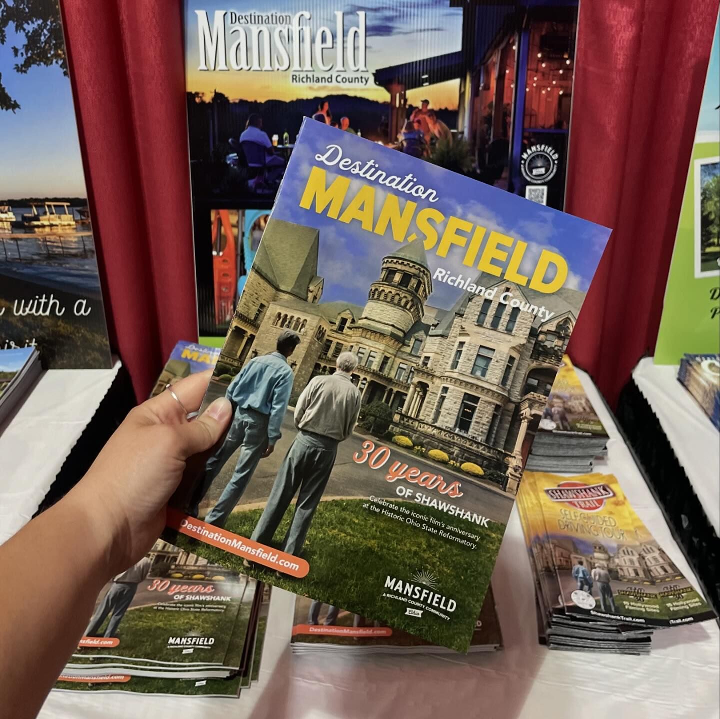 Hand holding Mansfield brochure with historic buildings and two men in suits, displayed on a table with other brochures.