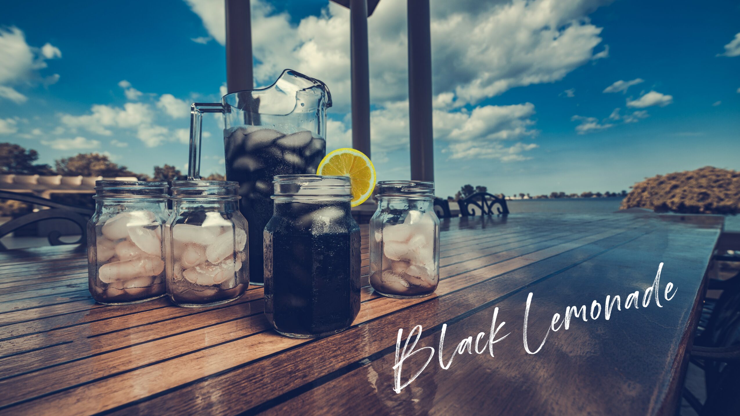 Glass jars and bottles with dark beverages and lemon slices on a wooden table outdoors, with a lake and sky in the background.