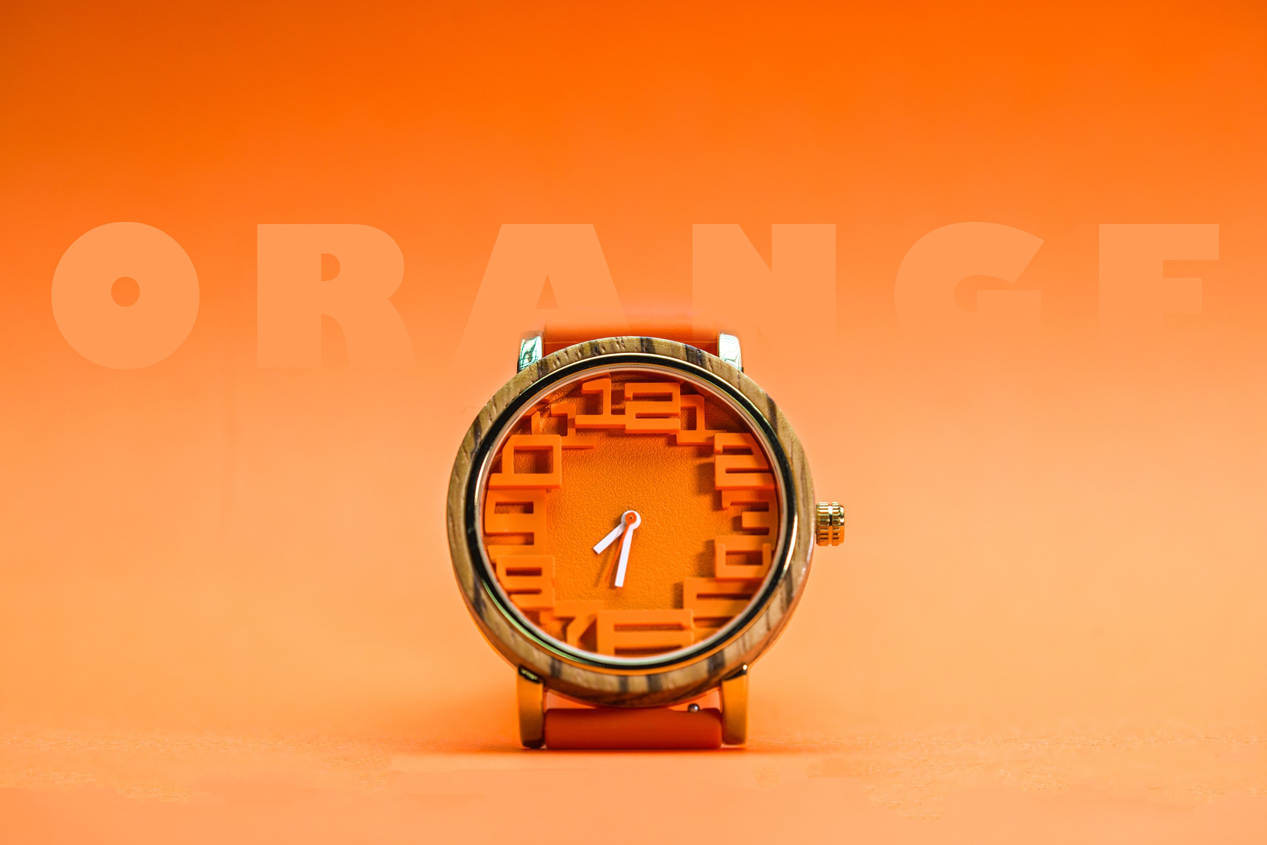 Watch with an orange face and gold band, displaying the time as 1:00, against an orange background with partially visible text.