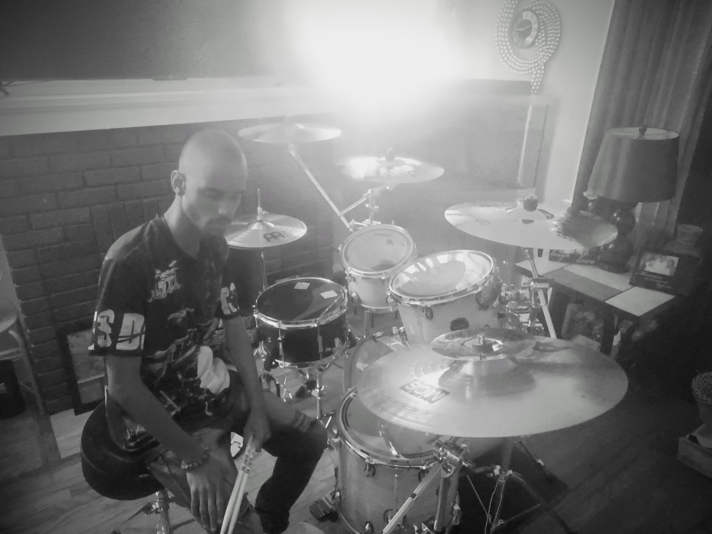 Person sitting near a drum set in a room with a lamp and window, smiling at the camera, black and white photo.