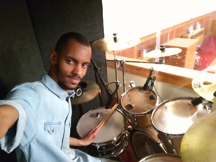 Young man taking a selfie with a drum set in a music studio, smiling, wearing a light blue shirt.