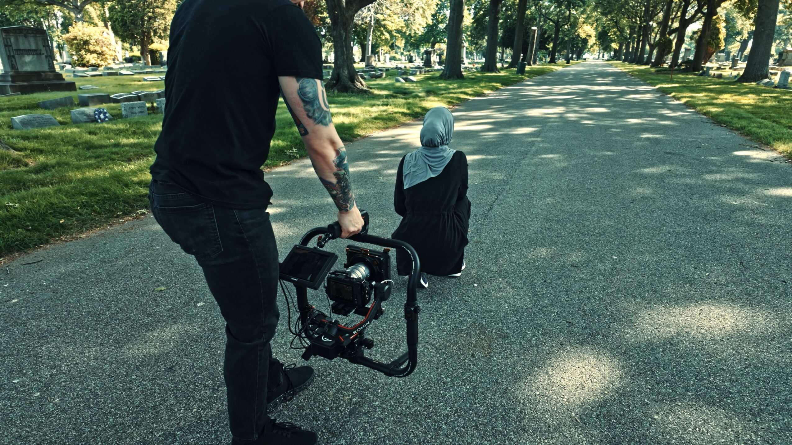 Person holding a camera on a gimbal filming a child sitting on a park road, trees and grass in background.