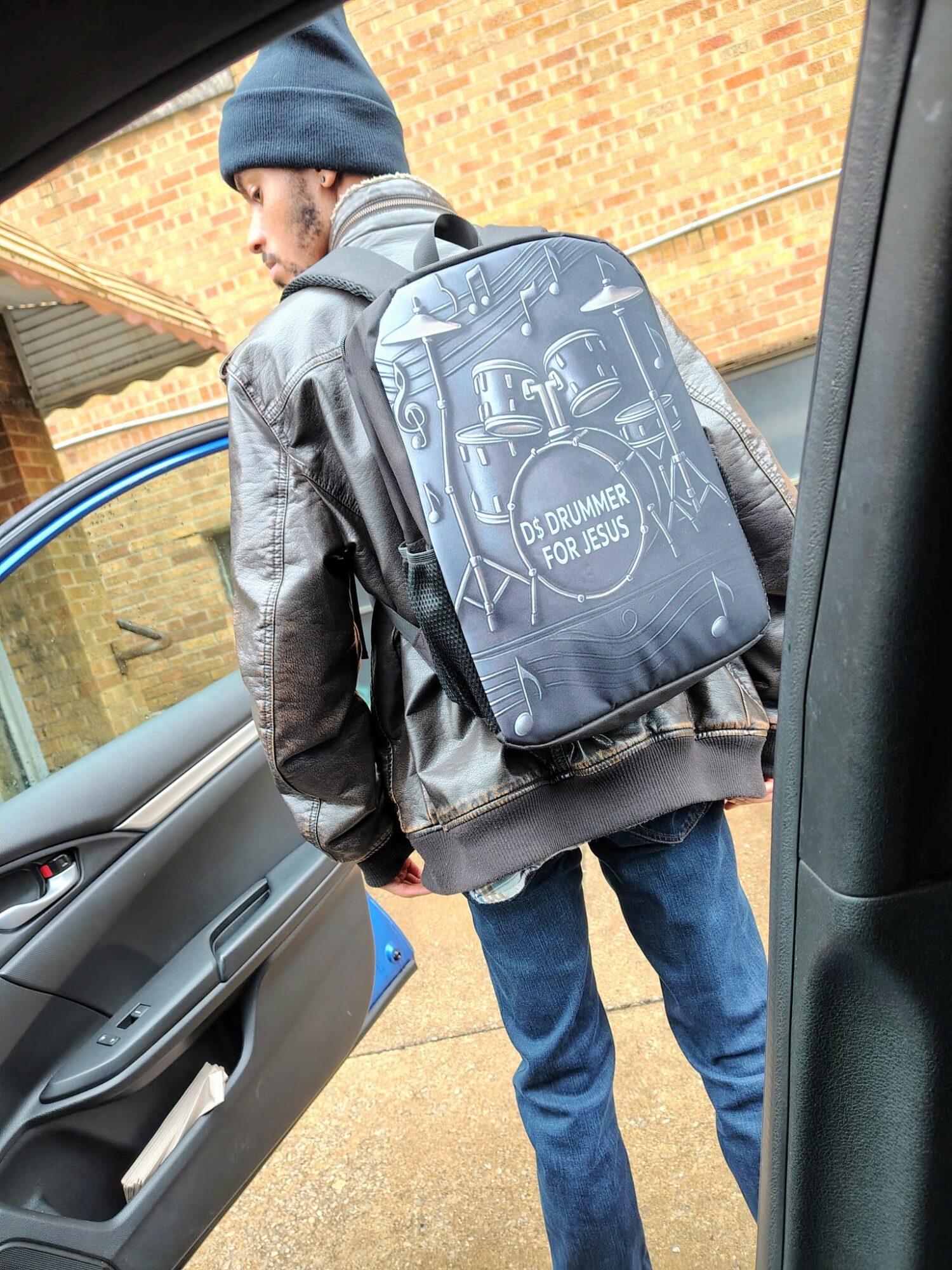 Person wearing a beanie and jacket with a backpack, standing outside near a vehicle, brick wall in background.