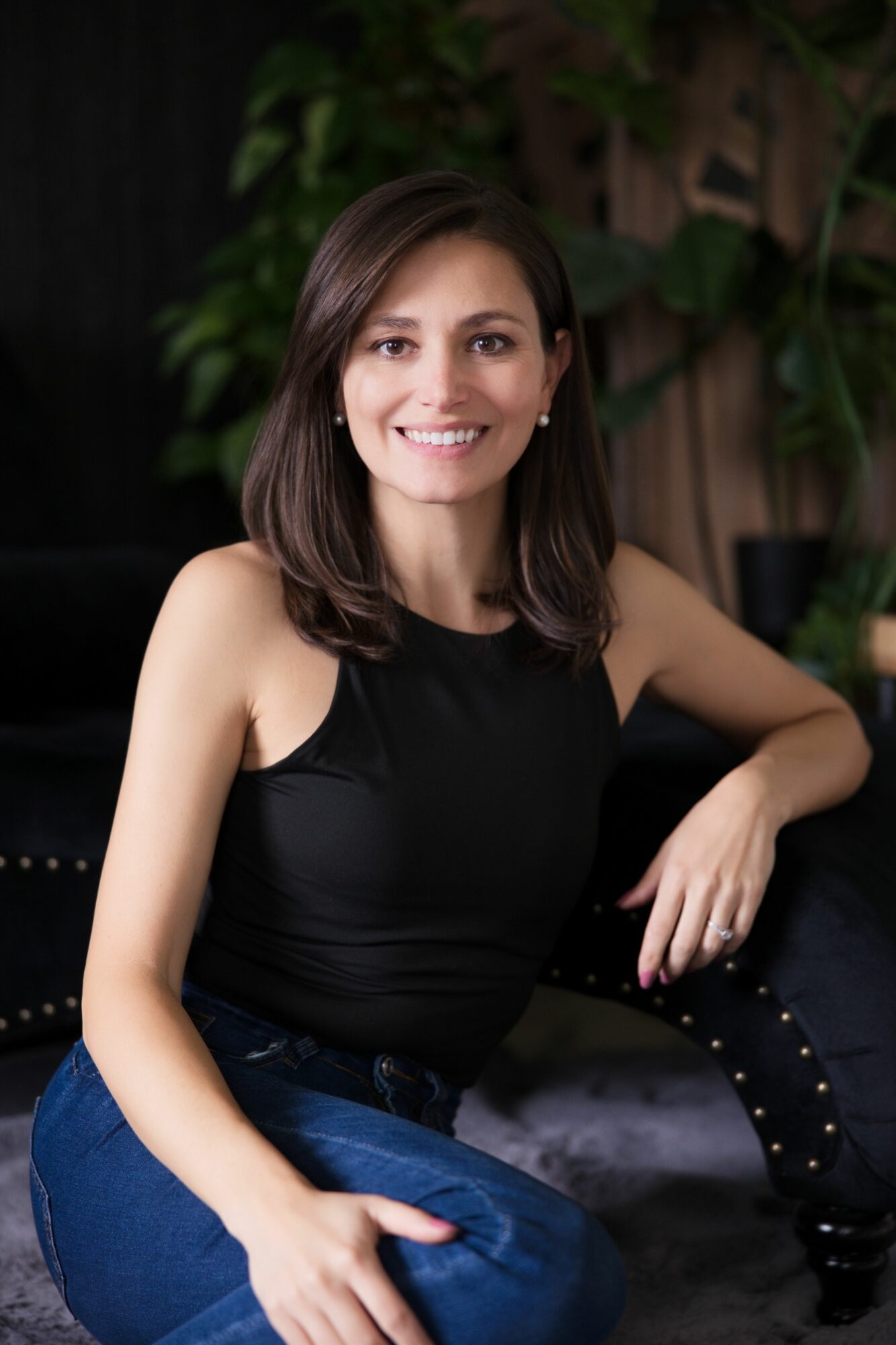 Woman with long brown hair smiling, wearing a black top and blue jeans, sitting outdoors with greenery in background.