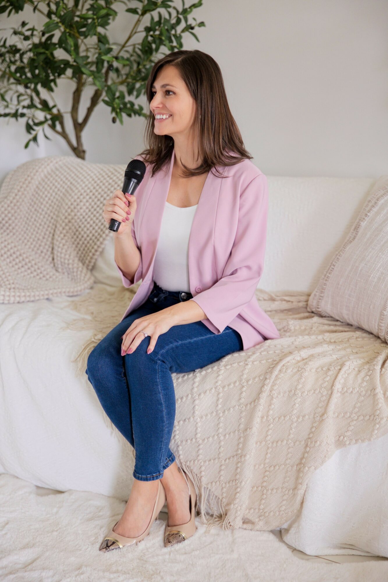 Woman sitting on a bed with a pink blazer, jeans, and high heels, holding a phone, in a room with a plant.