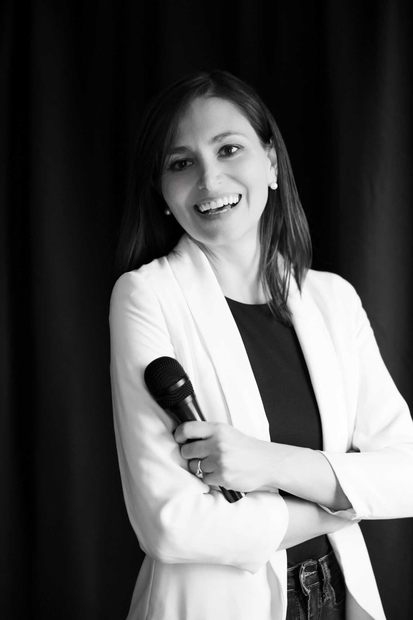 Woman smiling and holding a microphone, wearing a white blazer and black top, against a dark background.