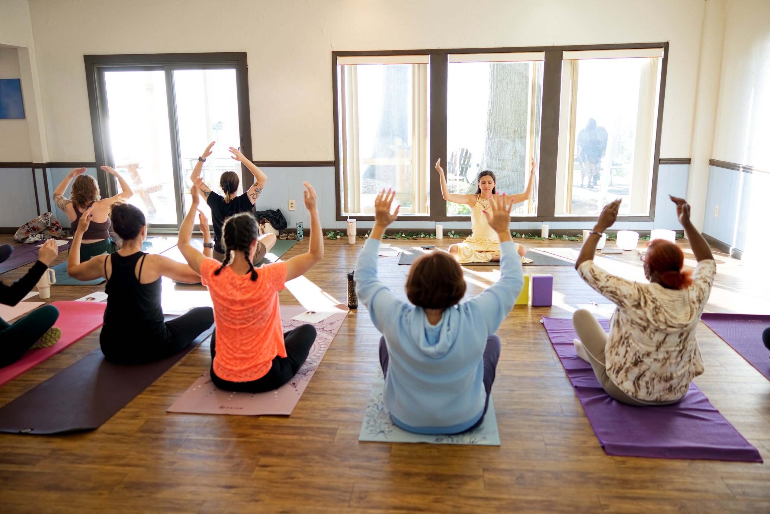 Group of people practicing yoga or meditation in a bright room with large windows, sitting on mats with hands raised.
